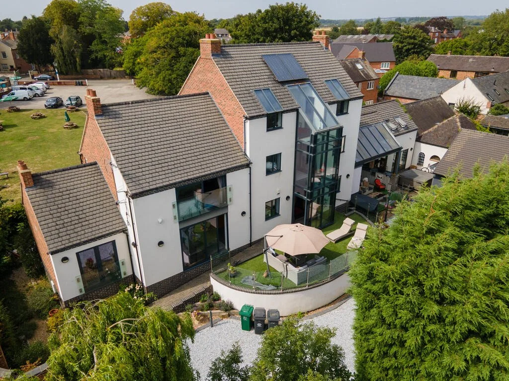 Merritt Estates. A modern multi-story house with a glass elevator shaft in the backyard, outdoor patio furniture with umbrellas, surrounded by trees and neighbouring homes.