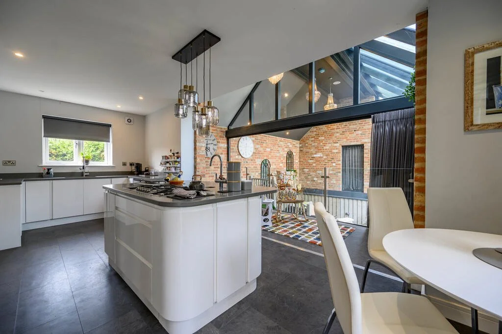 Merritt Estates. Modern kitchen with white cabinets, black countertop island, hanging pendant lights, and a dining area with white chairs. An open space with a brick wall and a balcony or loft area with a colourful rug and seating.