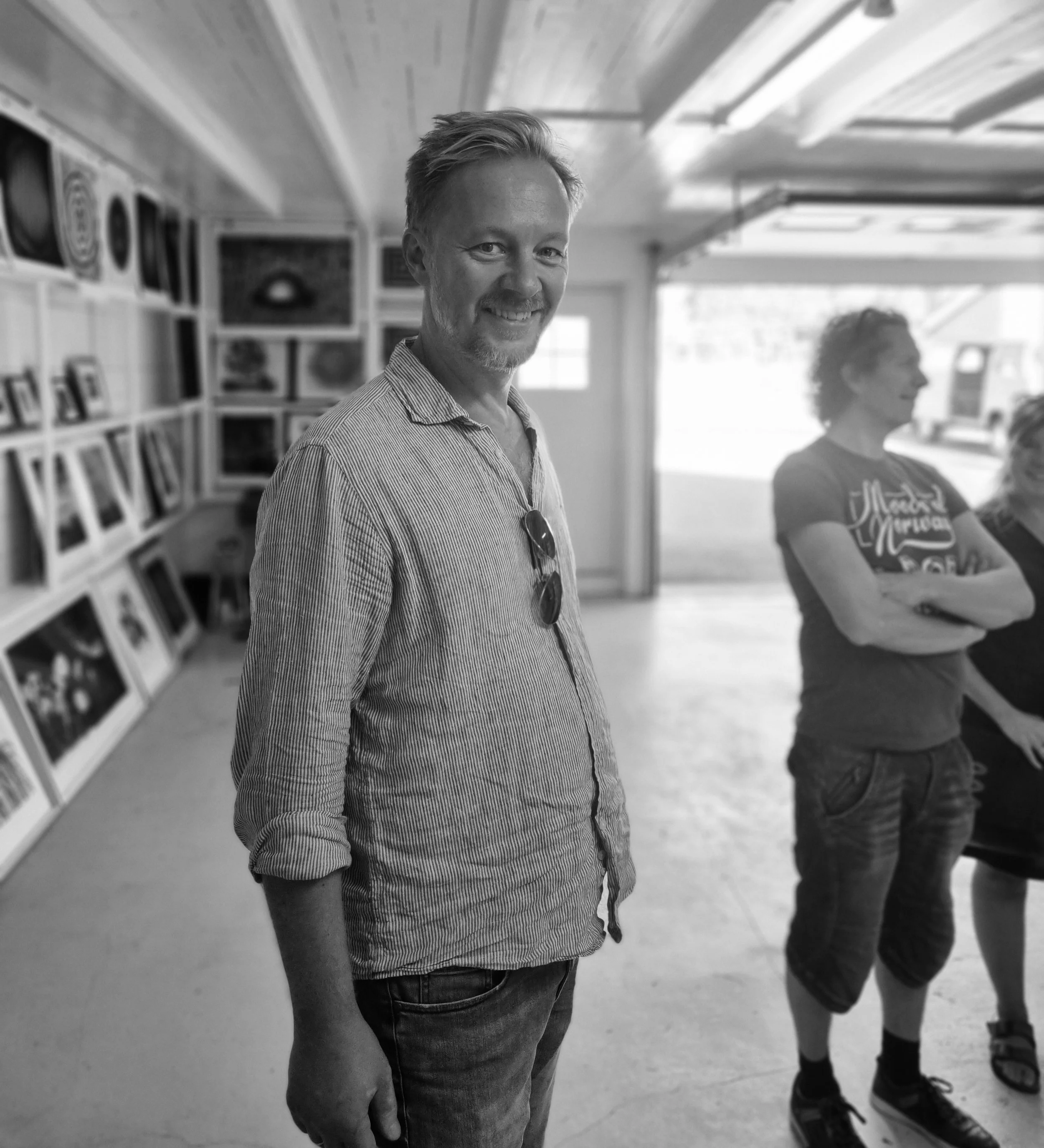En hvit-mann med kort, lysebrunt hår og skjegg, smiler til kamera, iført en stripete skjorte med ermer oppkuttet og solbriller hengende i halsen, står innendørs i et rom med bilder på veggen.