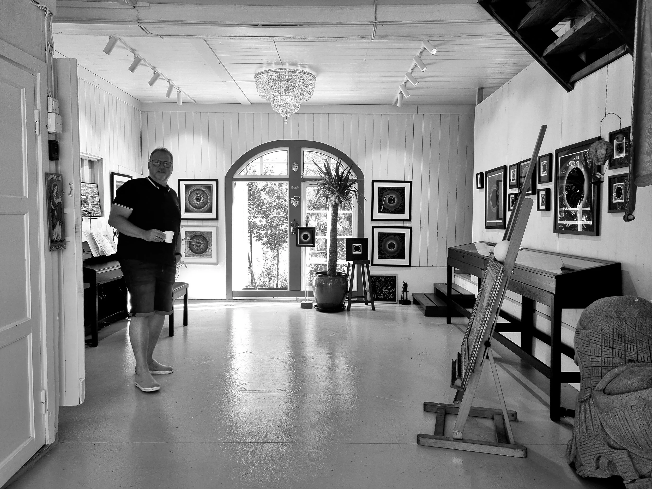 En mann med briller, svart T-skjorte og shorts står i en kunstutstilling med flere bilder på veggen, holder en kopp, i rom med hvite panelvegger og store vinduer.