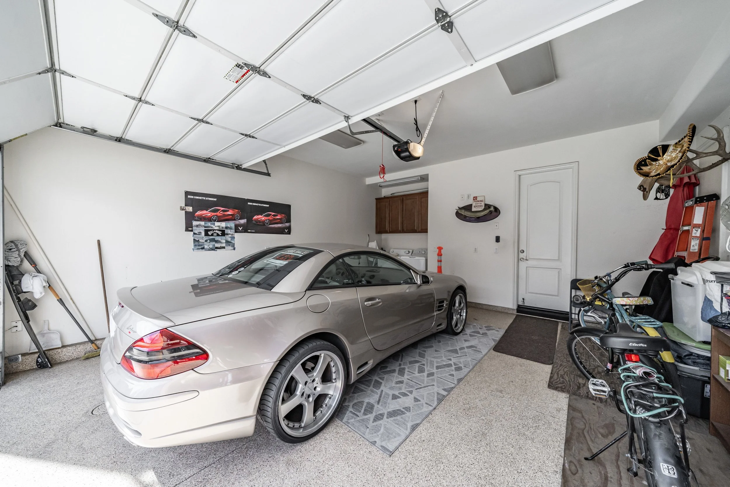 A garage with a silver sports car parked inside, various tools and bikes, a garage door partially open, and posters of red sports cars on the wall.