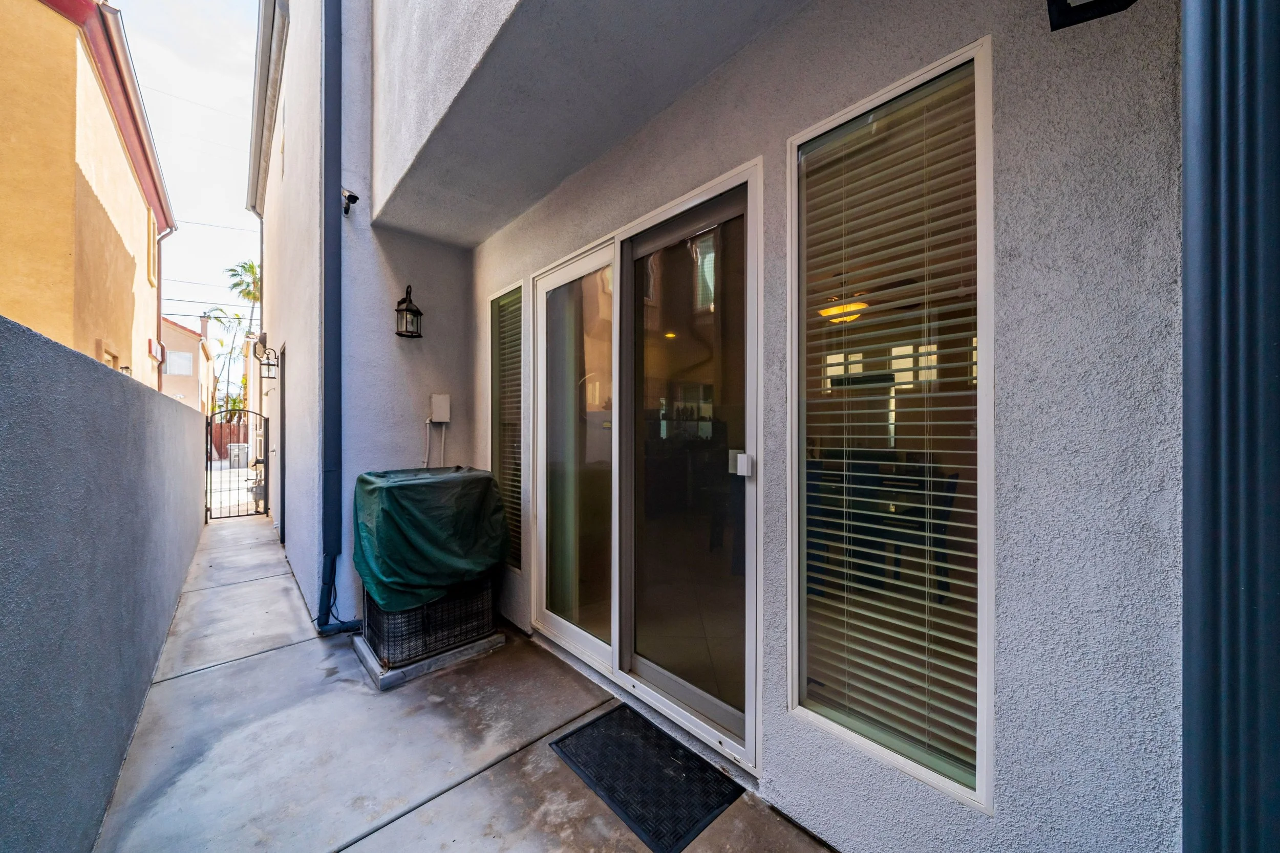 Backyard patio with sliding glass door, windows with blinds, potted plants, and ambient lighting fixtures on stucco wall.