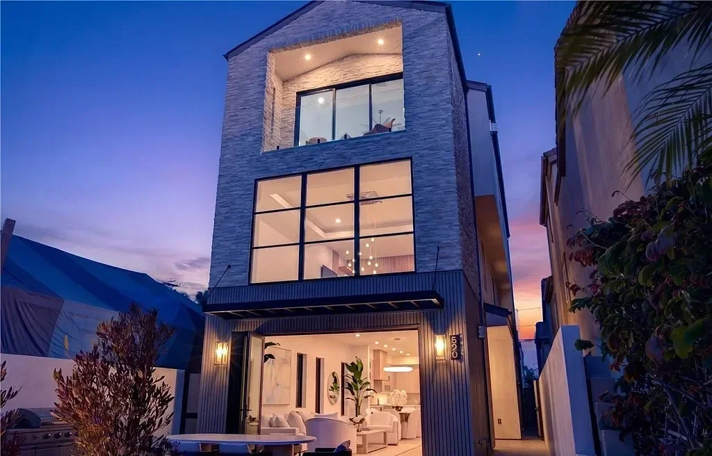 A modern three-story house with large glass windows in the evening, illuminated interior, and a patio area with outdoor seating.