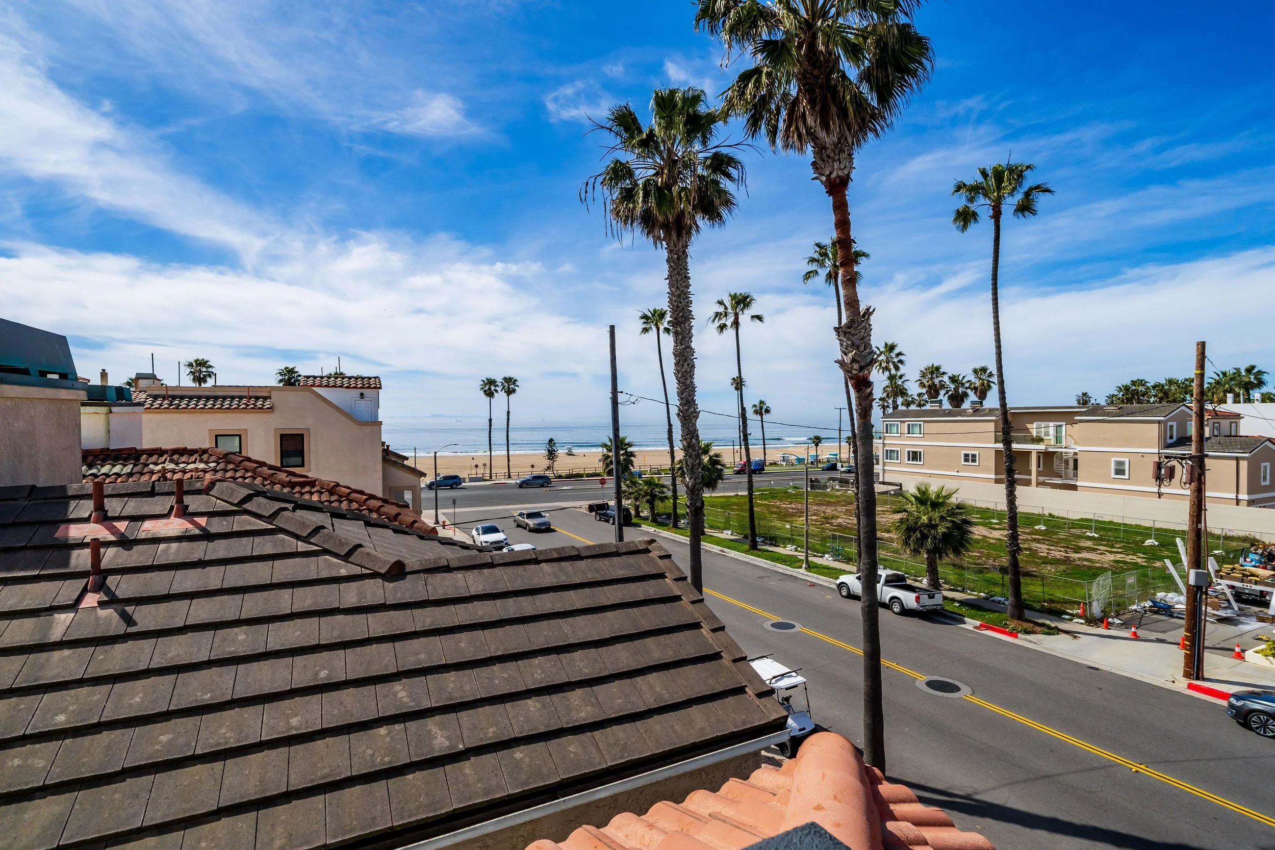 View of a street lined with palm trees, buildings, and parked cars with the ocean and a sandy beach in the background, under a blue sky with scattered clouds.