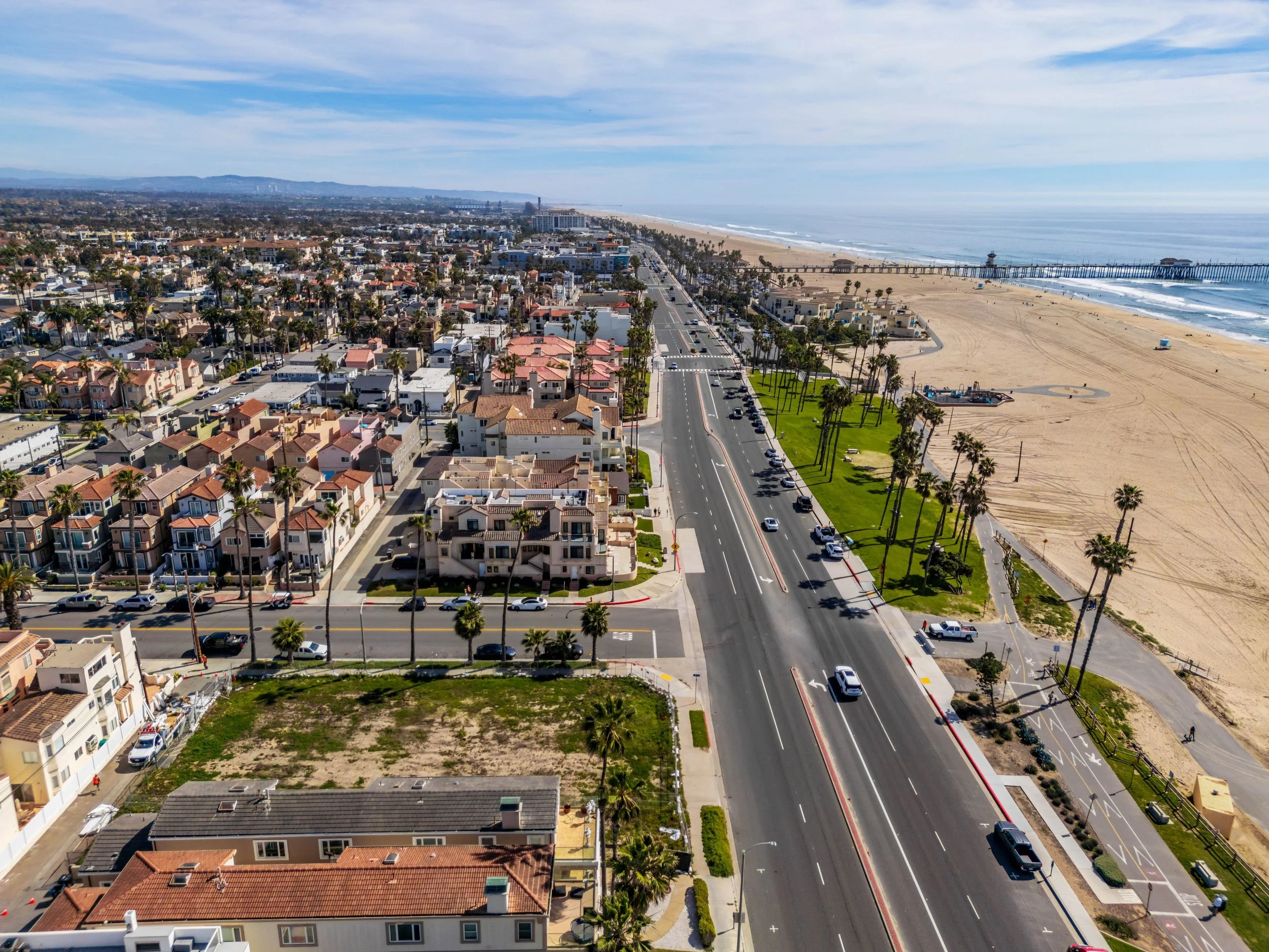 Aerial view of a coastal city with residential buildings, a main road lined with palm trees, and a sandy beach with a pier extending into the ocean in the background.