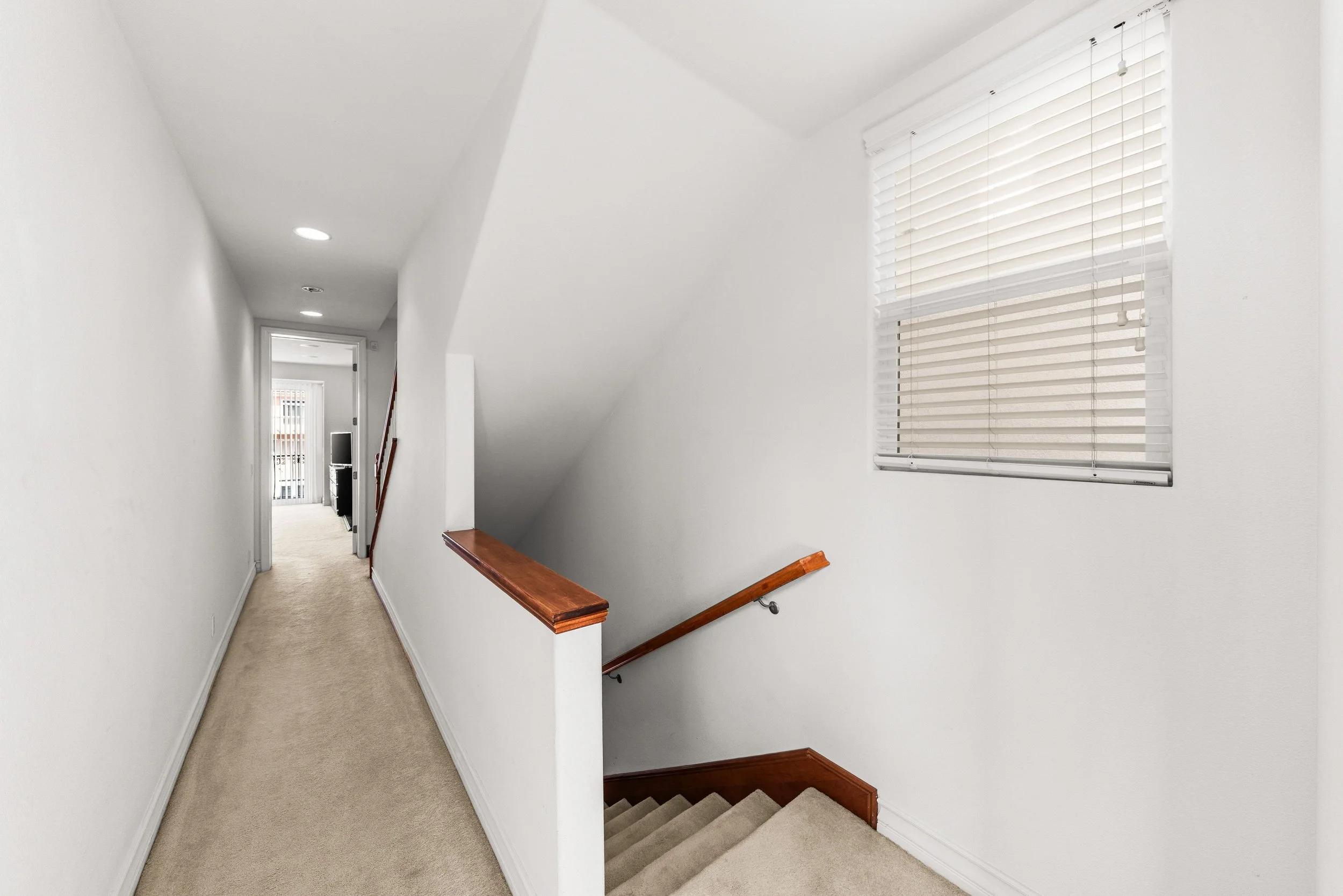 Interior view of a hallway with white walls, beige carpet flooring, a staircase with wooden handrails, and windows with closed blinds.