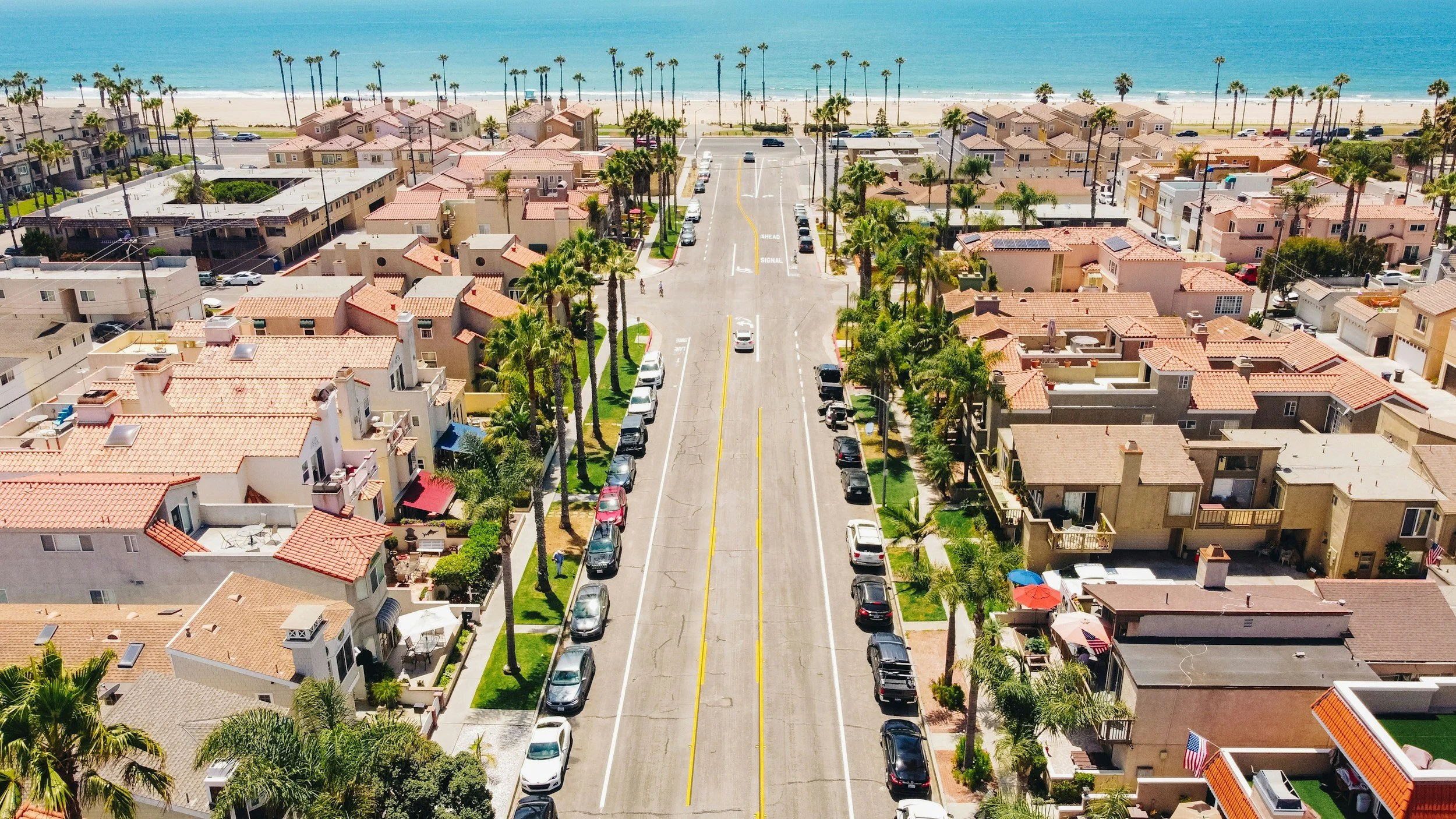 An aerial view of a beachside neighborhood with houses, parked cars, and palm trees, leading to a sandy beach and ocean.
