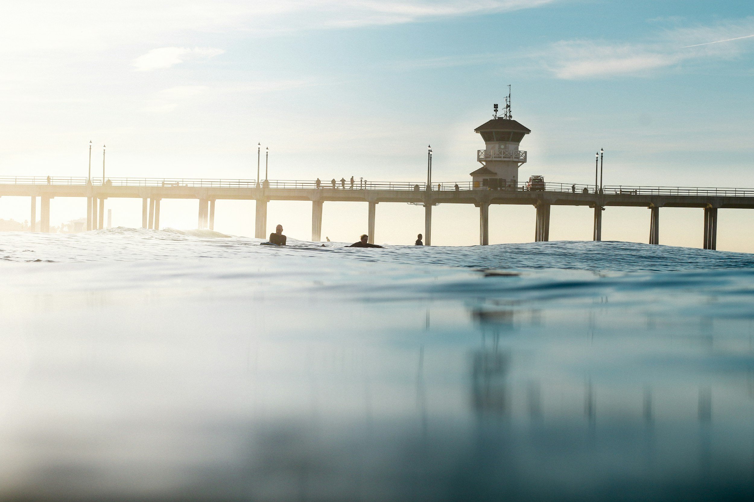 Huntington Beach pier extending over the ocean with people walking on it and a small watchtower at the end, under a sky with light clouds and contrail, during either sunrise or sunset.