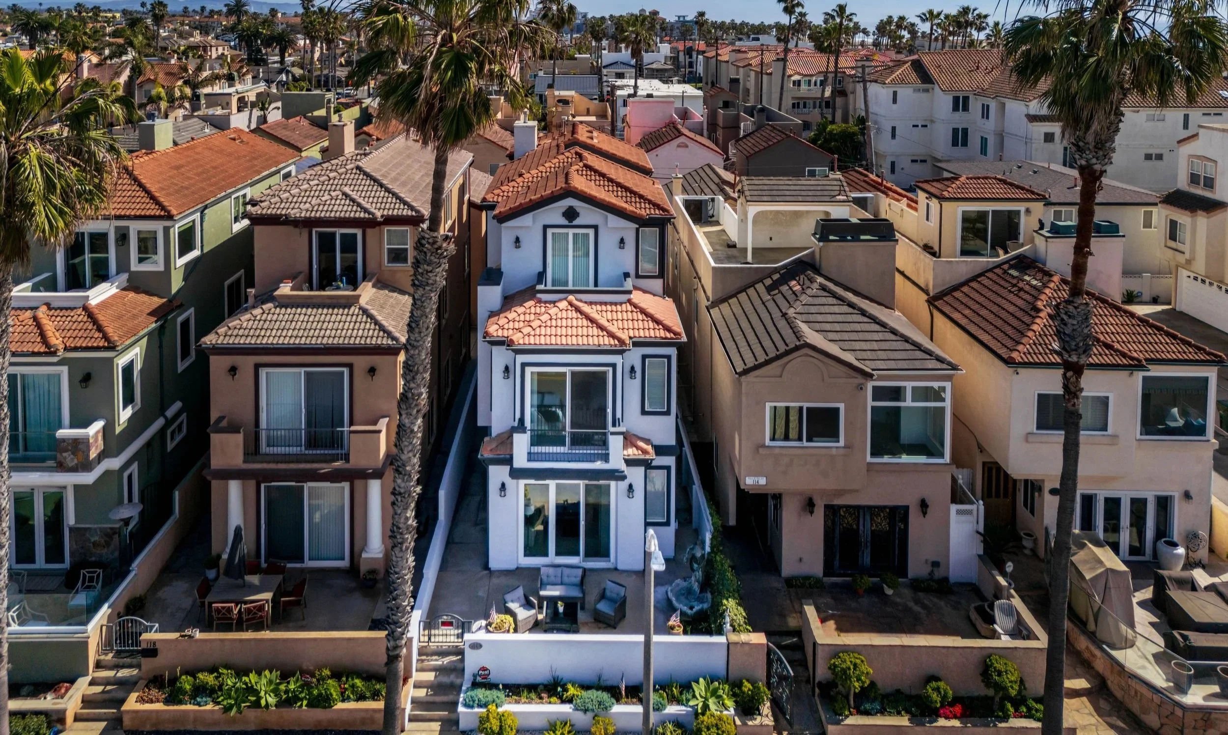 Aerial view of colorful suburban houses with tile roofs, palm trees, and well-maintained gardens in a sunny neighborhood.