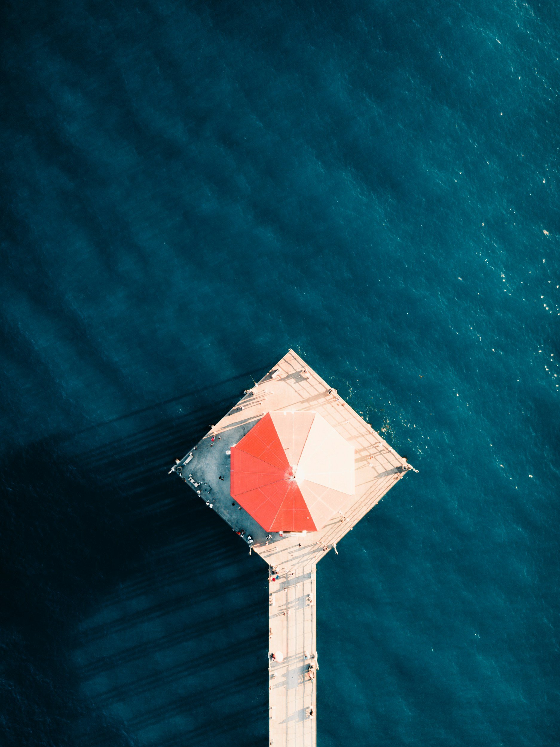 Aerial view of a pier extending into the ocean, with a large red and white rooftop structure at the end of the pier.