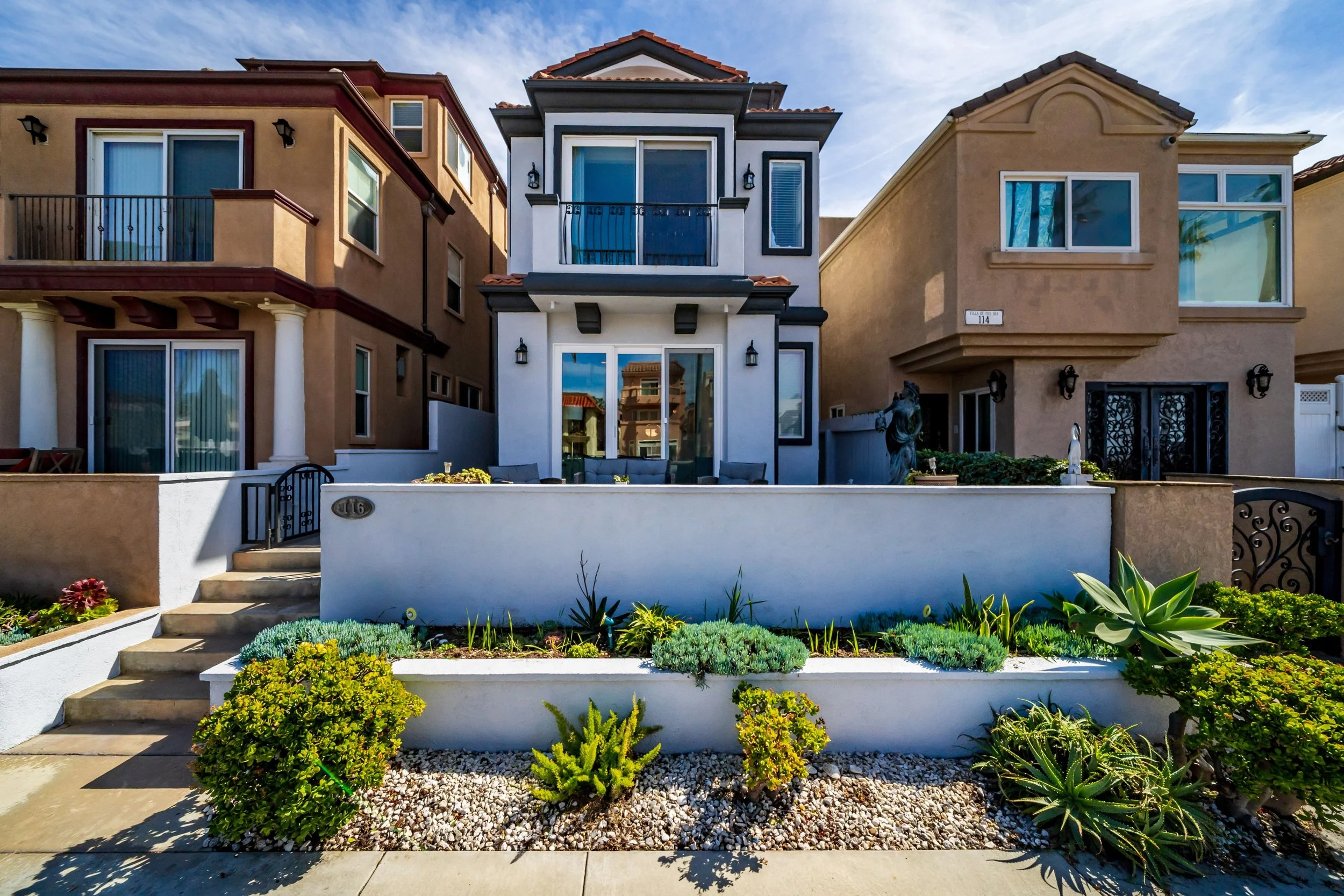 Front view of three modern houses with a garden and steps in the foreground under a blue sky with clouds.