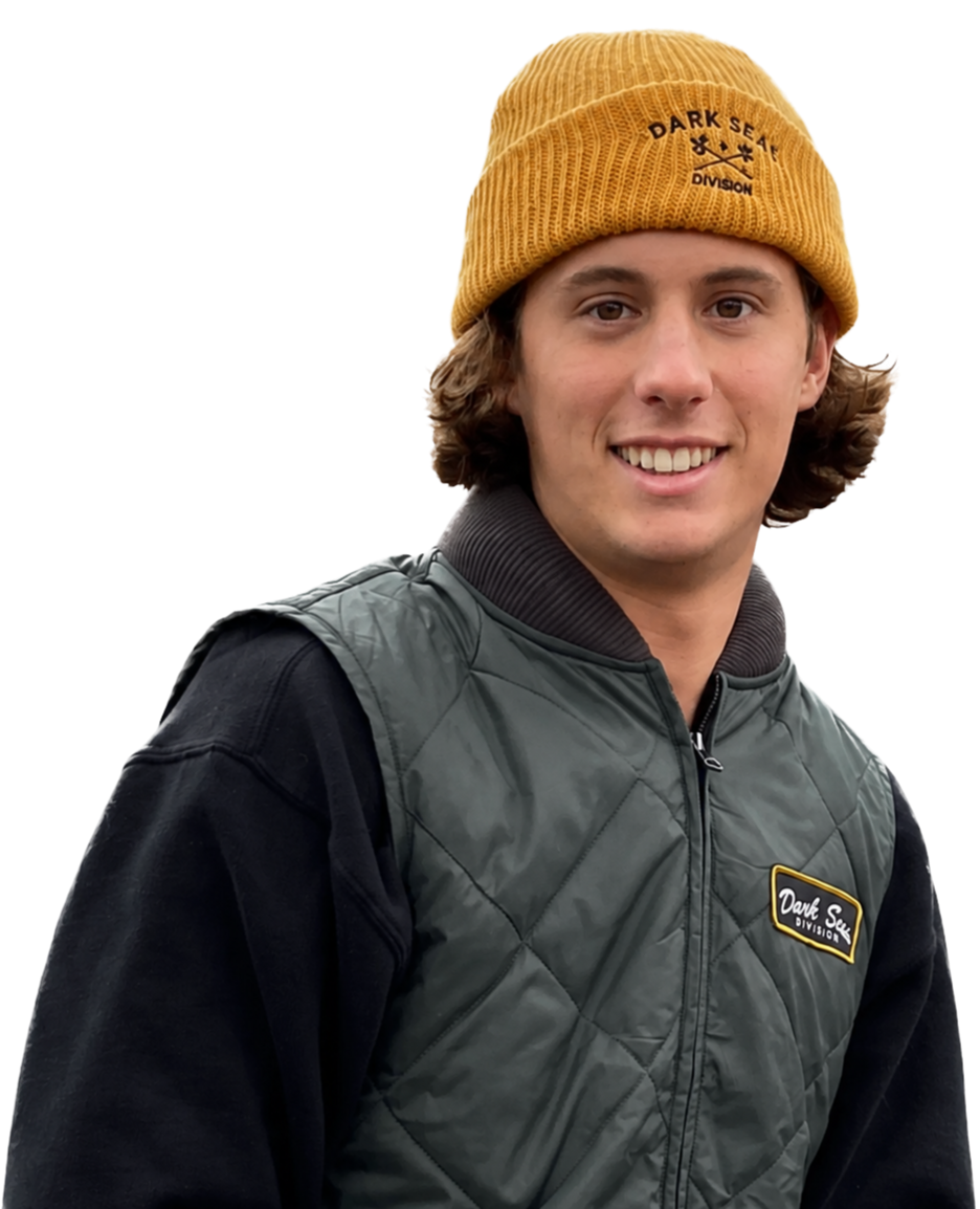 A young man smiling, wearing a yellow beanie with 'Dark Sea Division' logo and a dark green quilted vest over a black hoodie.