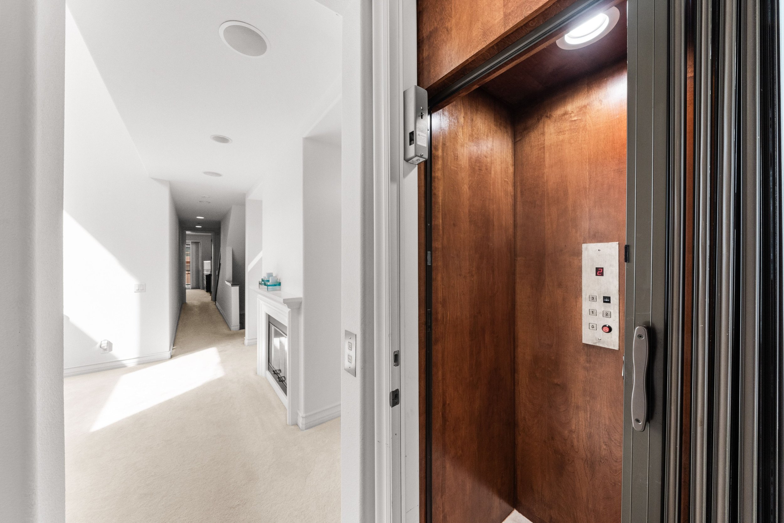 View from inside an elevator showing the elevator doors opening to a hallway with white walls and a beige carpeted floor.