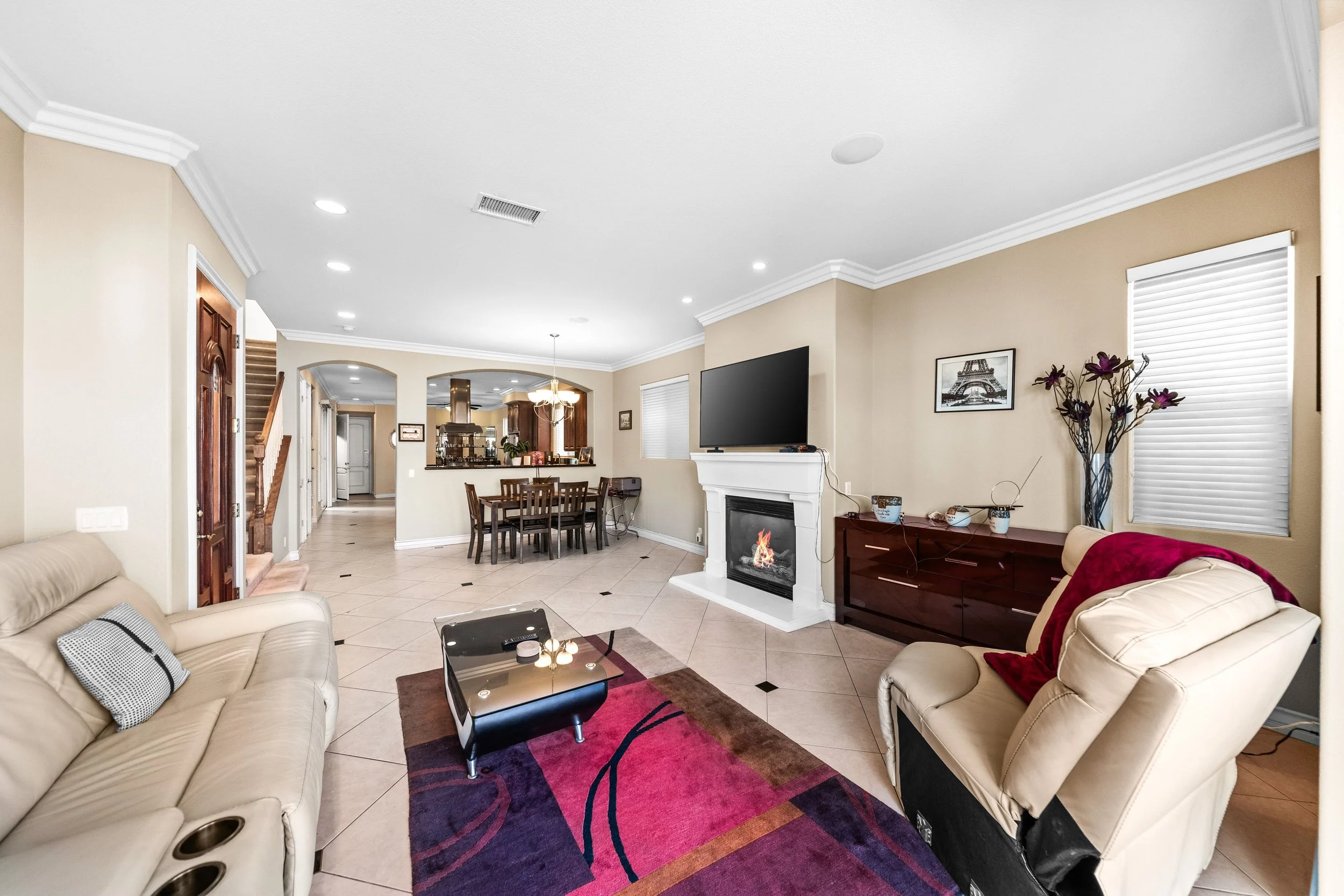 A spacious living room with beige walls, a white ceiling, tiled floor, and various furniture including a beige leather couch, a matching armchair with a red blanket, and a wooden coffee table with a purple and red rug underneath. There is a fireplace