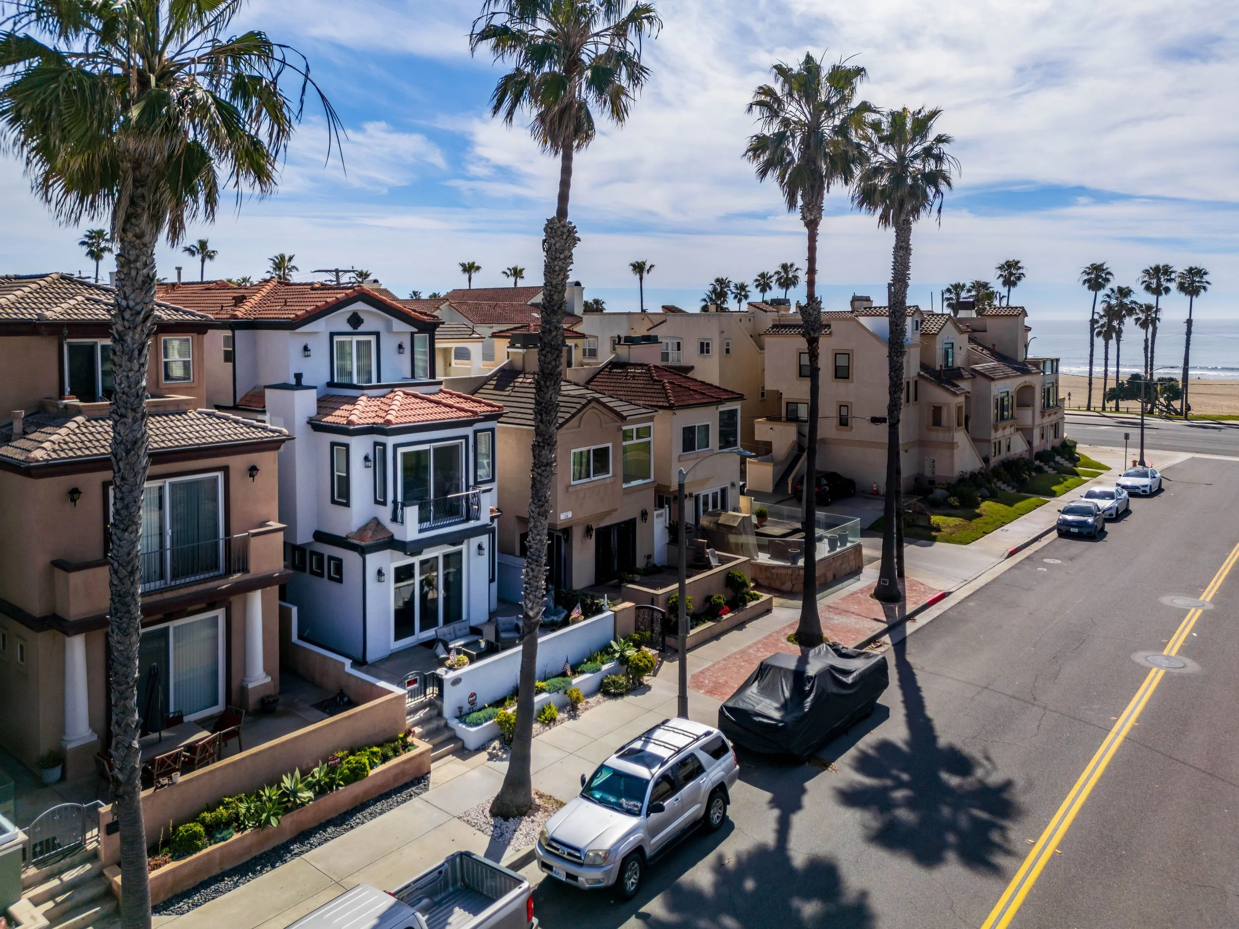A sunny coastal street with colorful houses, palm trees lining the sidewalk, parked cars, and a view of the beach and ocean in the distance under a partly cloudy sky.