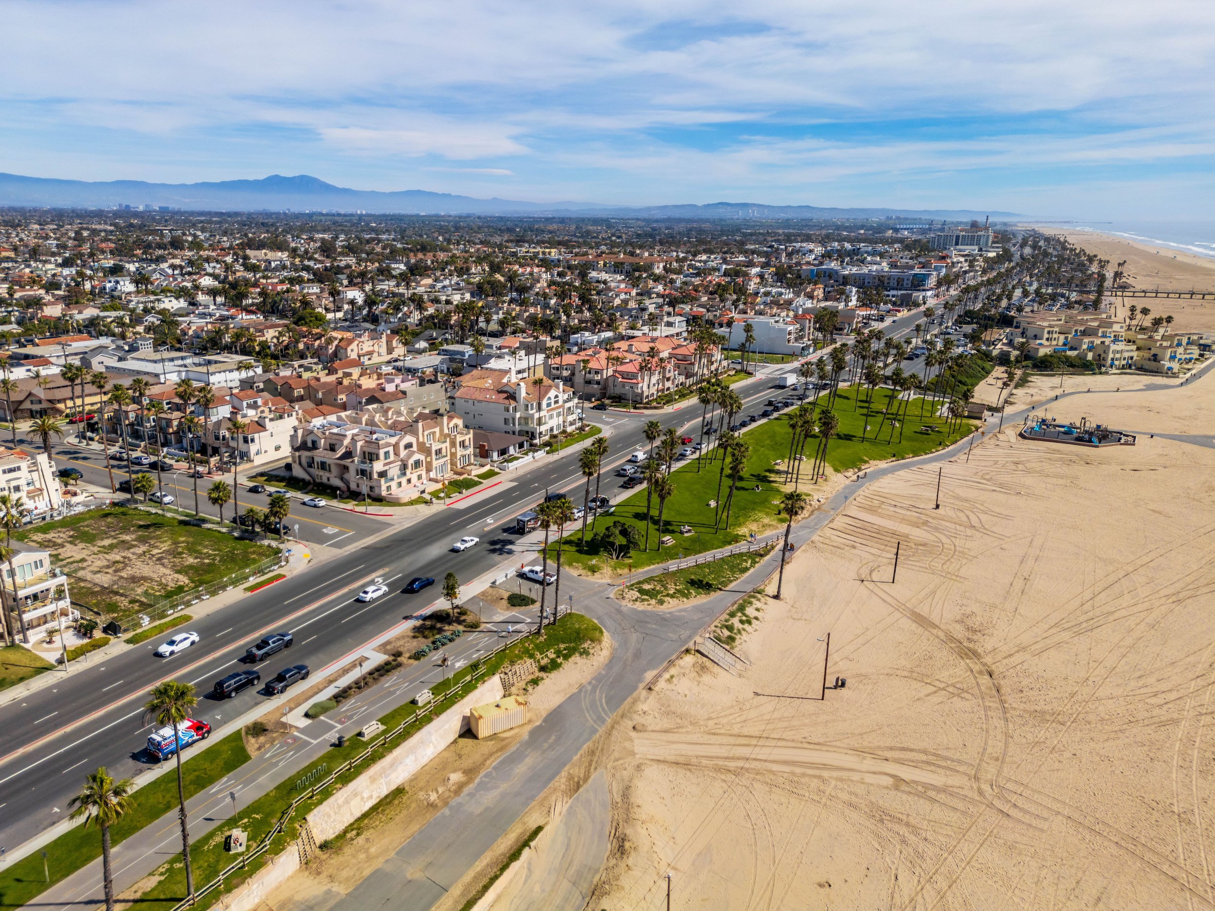 Aerial view of a coastal city with residential buildings, palm trees, a beach, and the ocean in the background on a partly cloudy day.