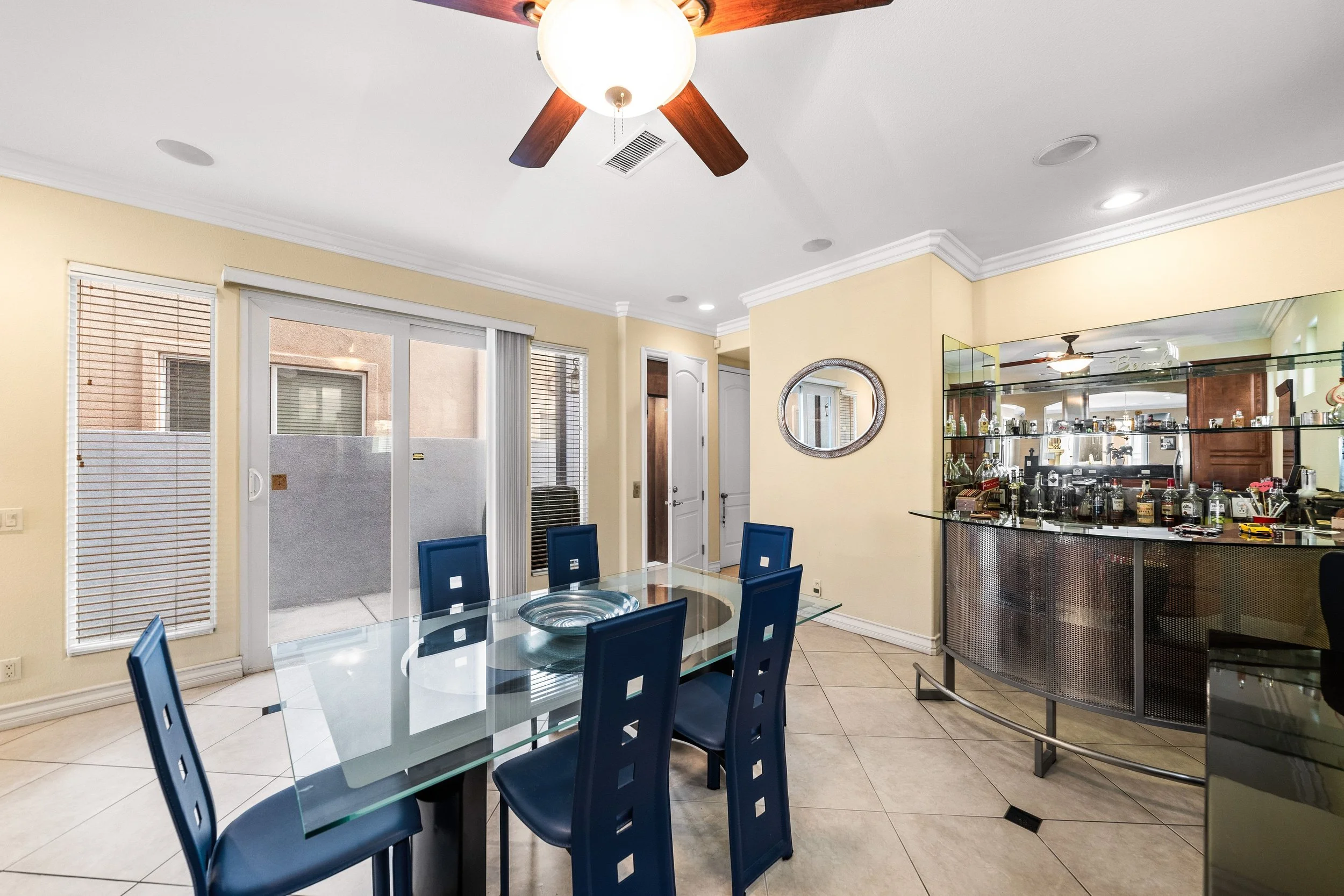 Dining area with a glass table surrounded by blue chairs, a ceiling fan with wooden blades, sliding glass door with vertical blinds, a wall mirror, a bar counter with bottles, and windows with blinds.