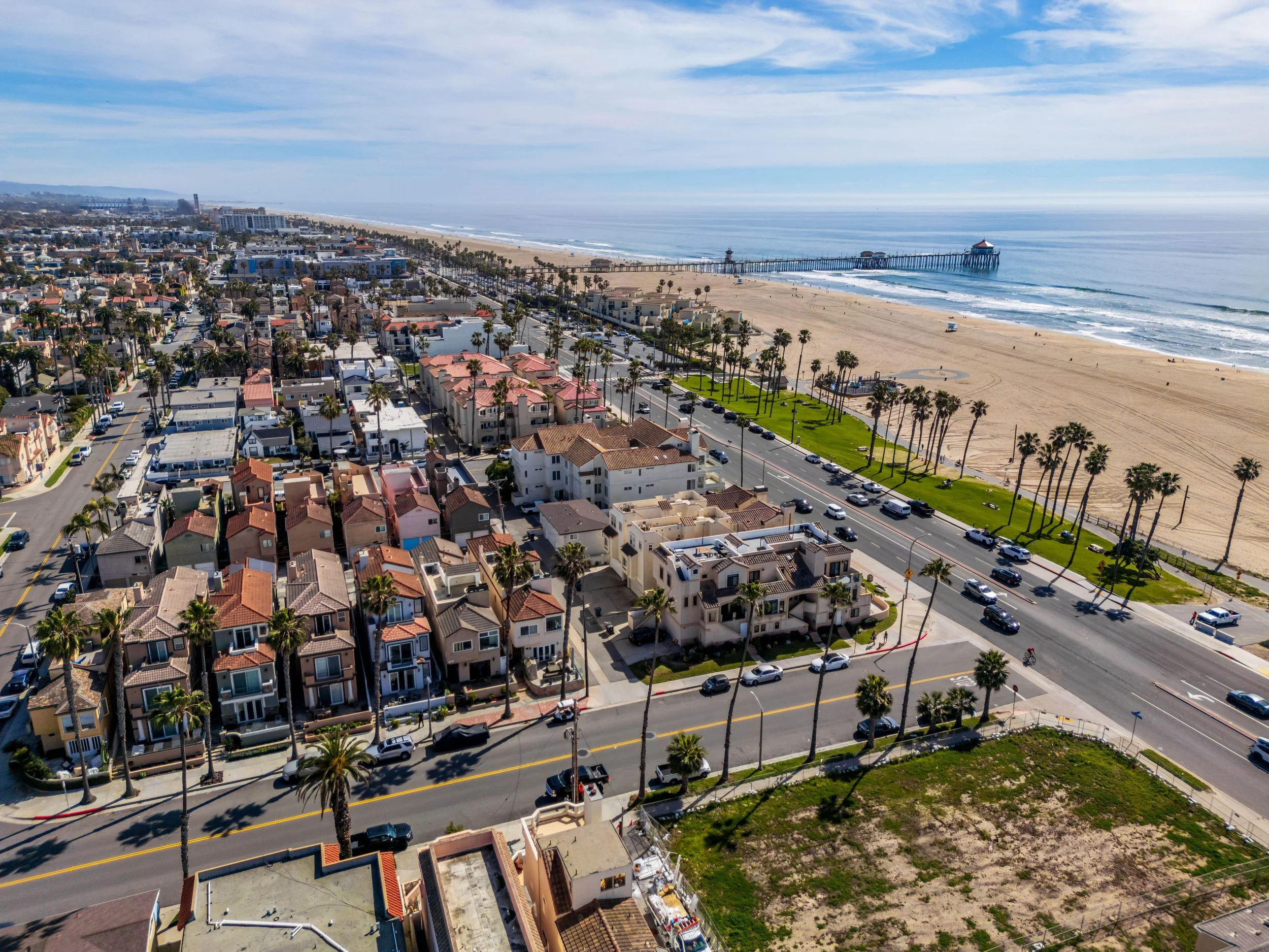 Aerial view of a coastal cityscape with residential buildings, palm trees, a beach, and a pier extending into the ocean under a partly cloudy sky.