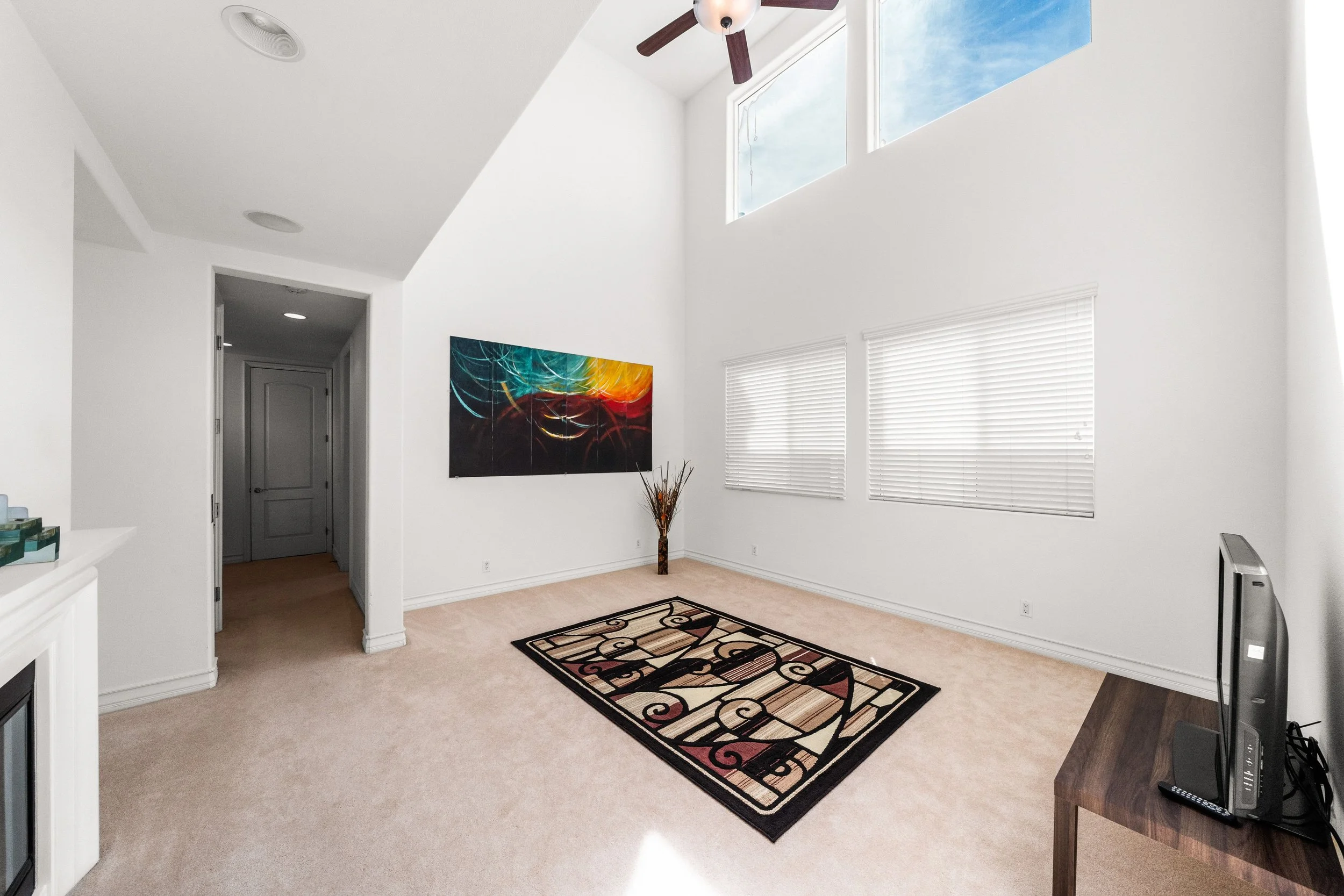 Living room with white walls, high ceiling, ceiling fan, window with blinds, abstract painting, decorative floor rug, and wooden console table.