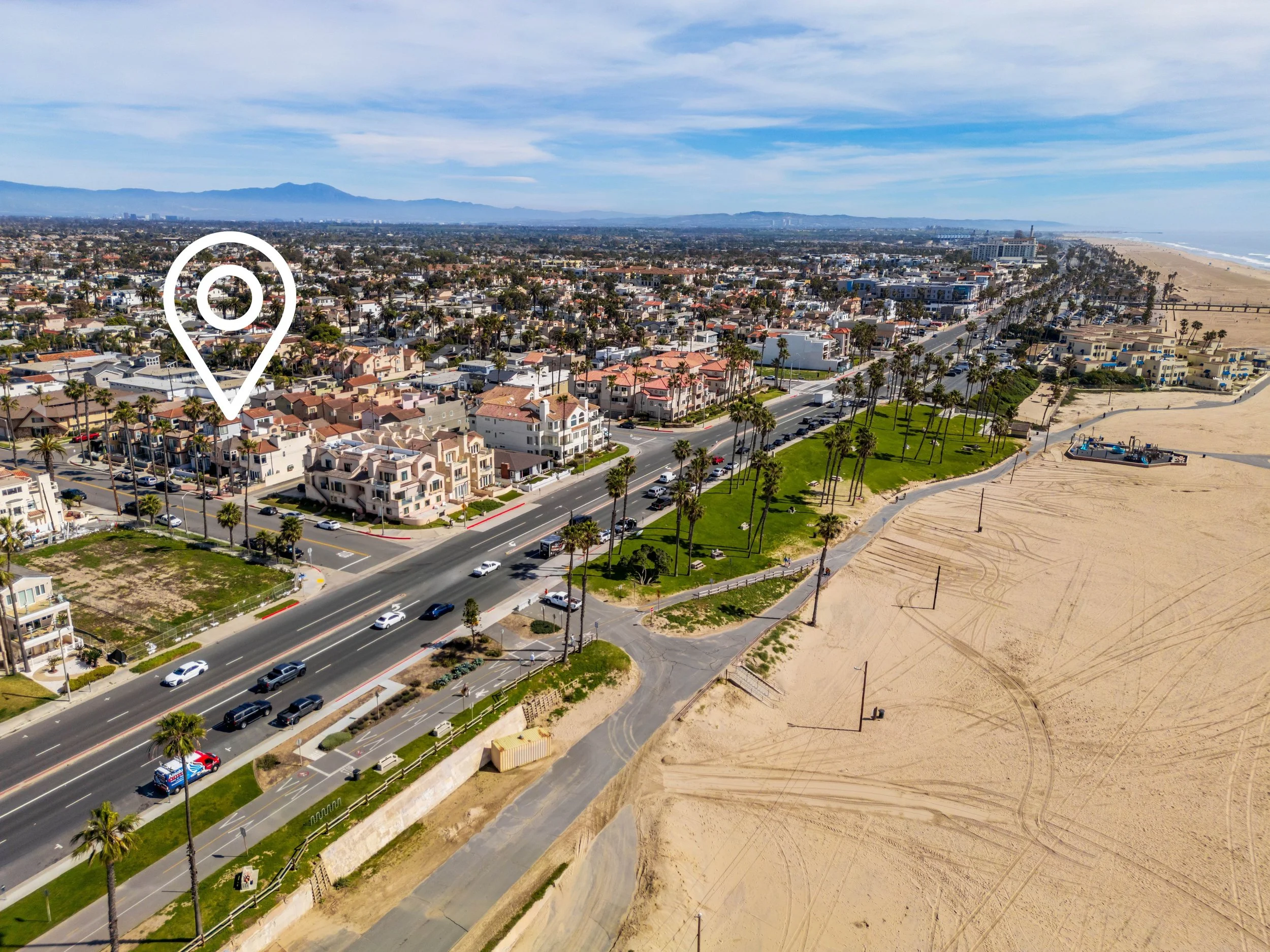 Aerial view of a coastal city with residential houses, palm trees, a beach, and the ocean in the background, with a marker indicating a specific location.