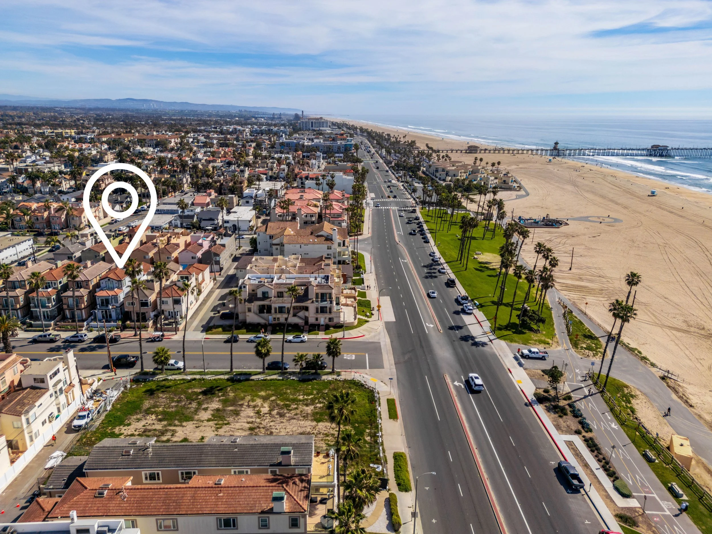 Aerial view of a coastal city with residential houses, a main road, and a long pier extending into the ocean, with beach and ocean waves in the background.