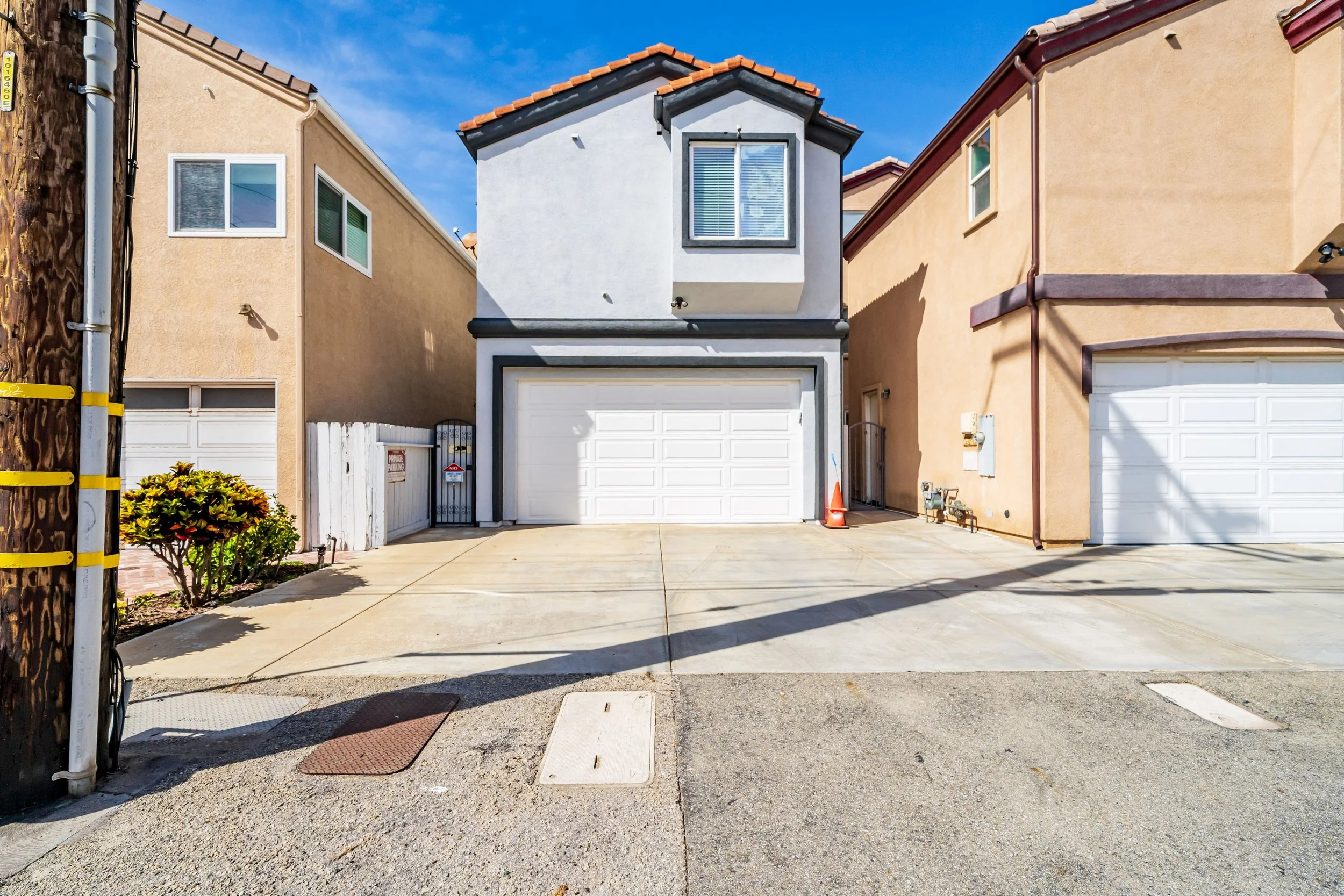A narrow white two-story house with a garage on the first floor and a window on the second floor, situated between two taller beige and orange houses. The driveway in front has two manhole covers and a small bush on the left, with a bright blue sky a