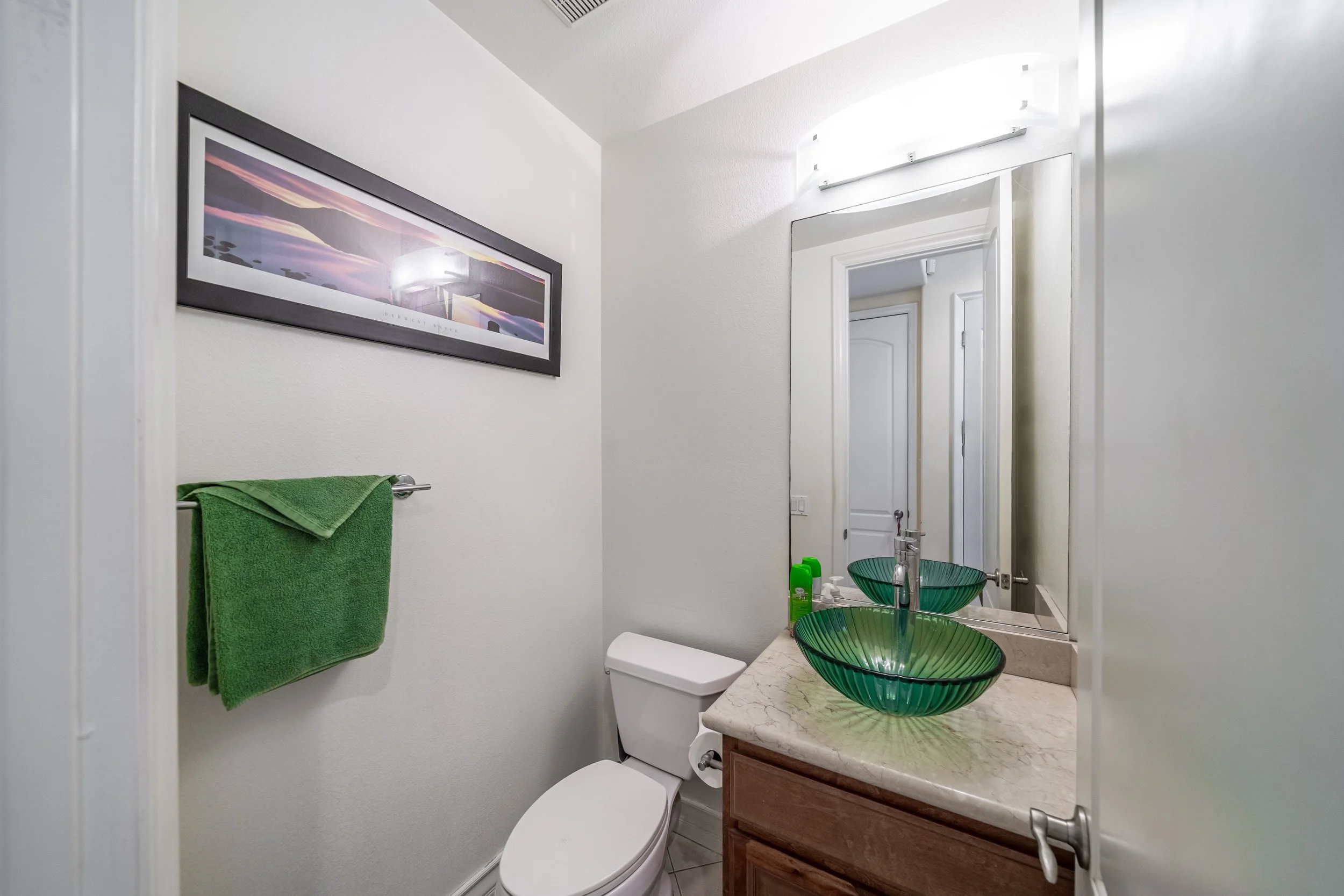 A small bathroom with a white toilet, green vessel sinks on a marble countertop, and a mirror. A framed landscape artwork hangs on the wall, and a green towel is on a towel rack. There are toiletries on the counter, and a door is partially visible.