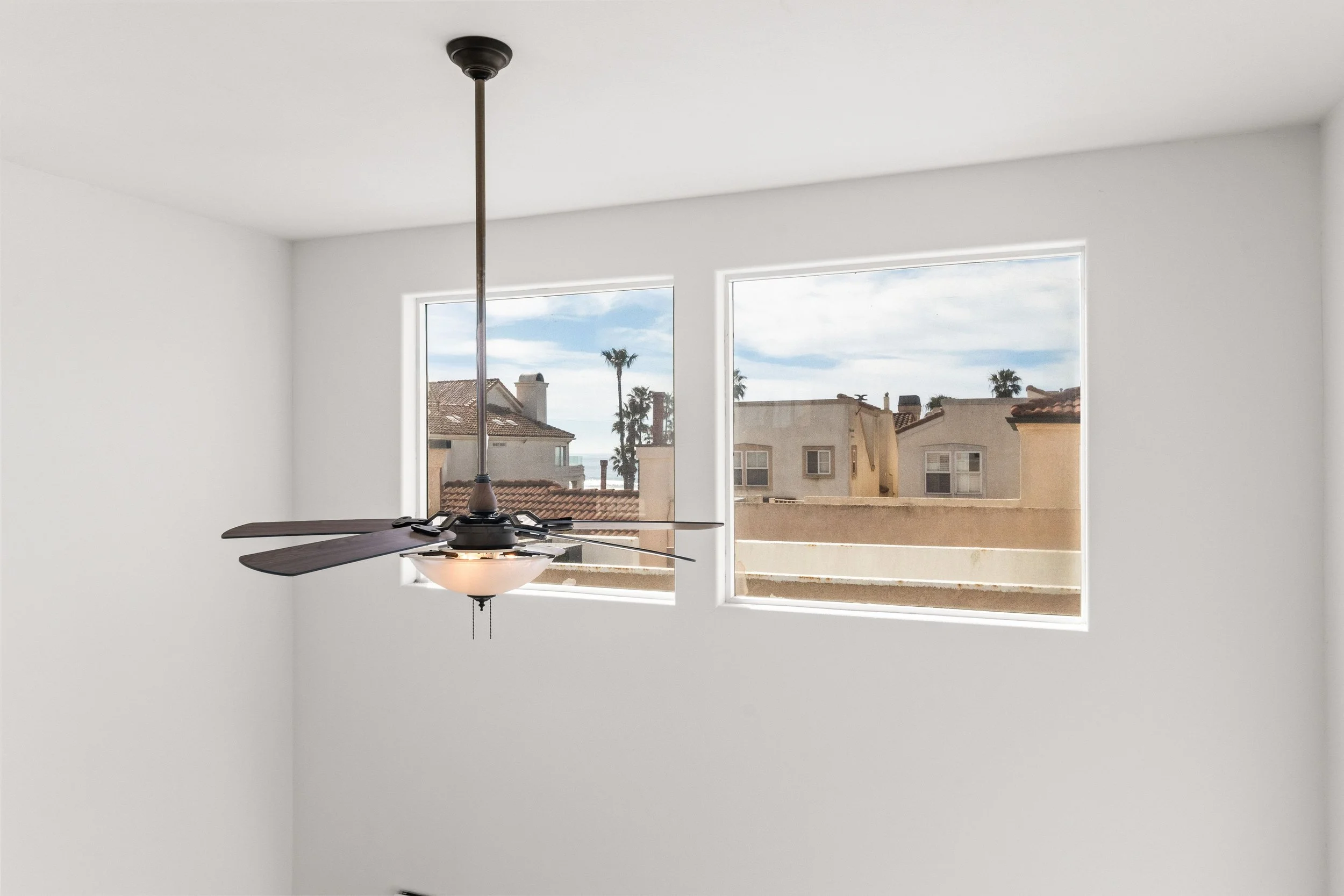 A room with white walls and two large windows with a view of neighboring houses and palm trees. A ceiling fan with a light fixture is hanging from the ceiling.
