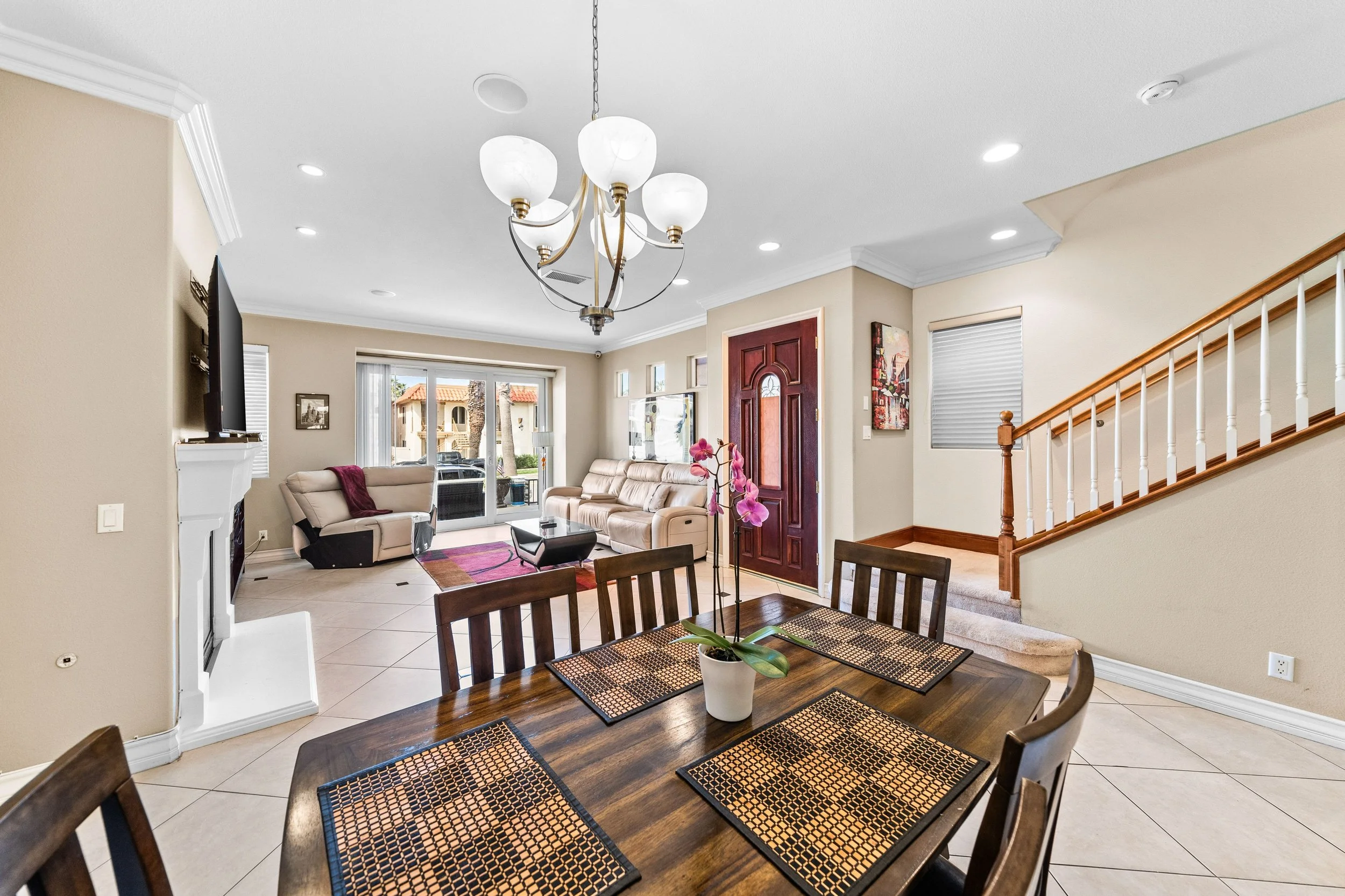 Bright living room with beige walls, a large beige sofa, a glass door leading outside, a staircase with wooden railing, a dining table with woven placemats and a potted orchid, and a ceiling chandelier with five white globes.