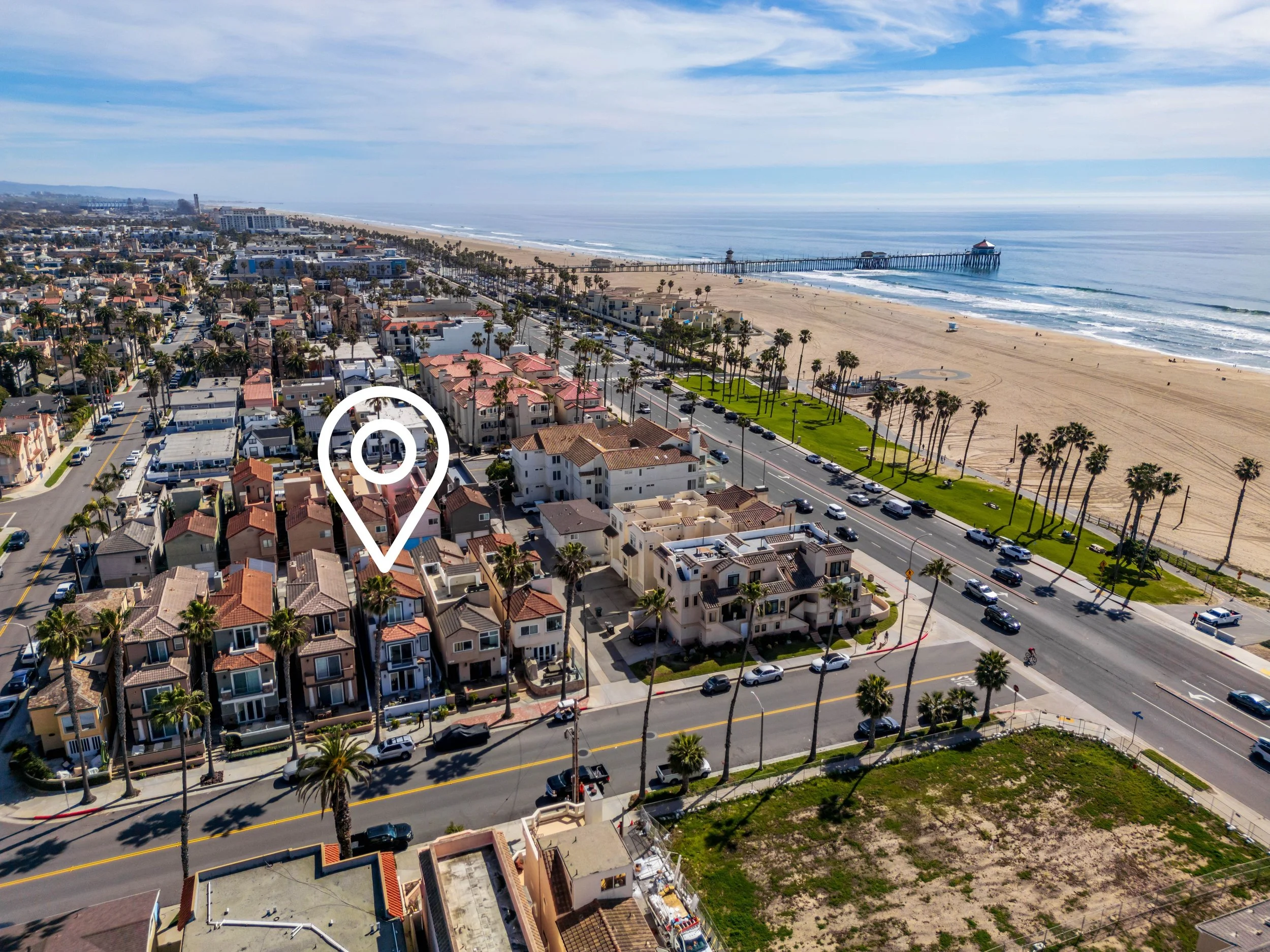 Aerial view of a coastal city with residential houses, palm trees, a beach, and a pier extending into the ocean under a partly cloudy sky.