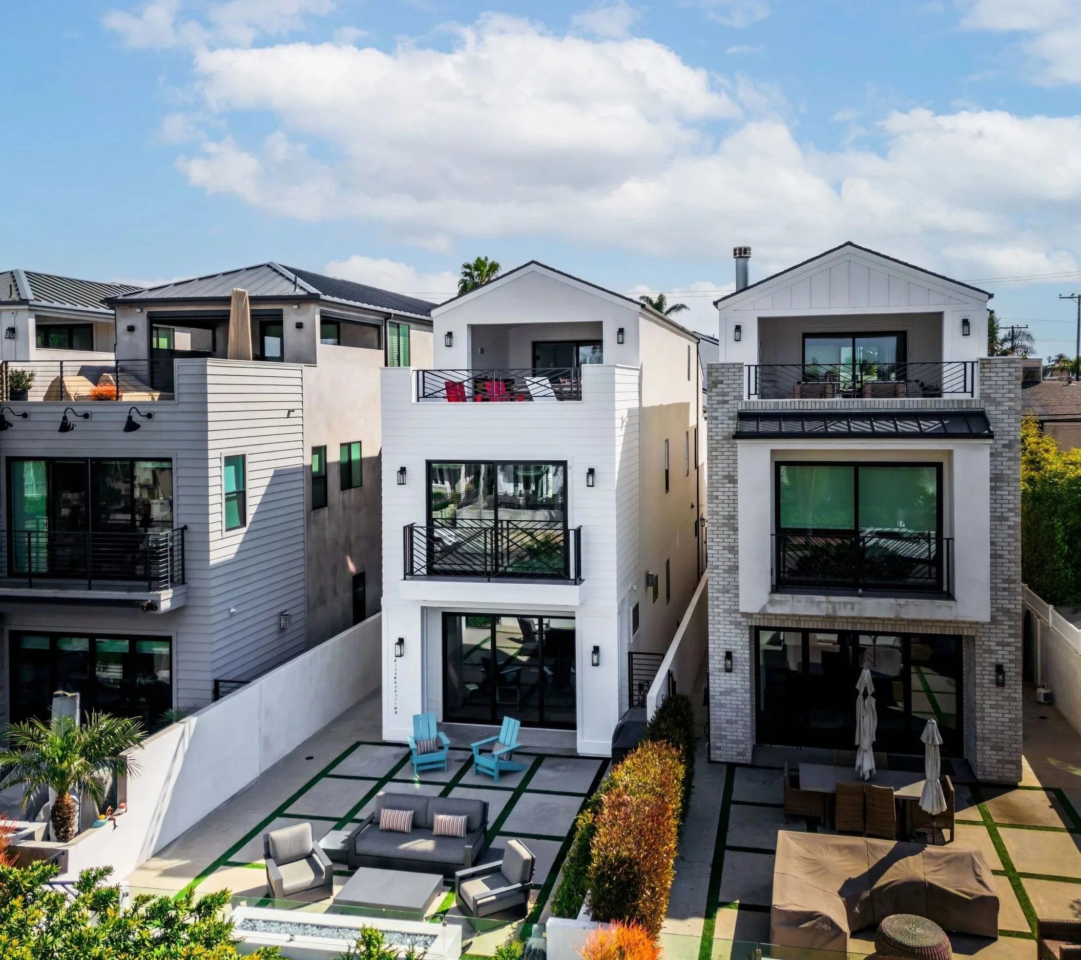 View of a modern apartment complex with balconies, outdoor seating area with chairs, umbrellas, and lush landscaping under a partly cloudy sky.
