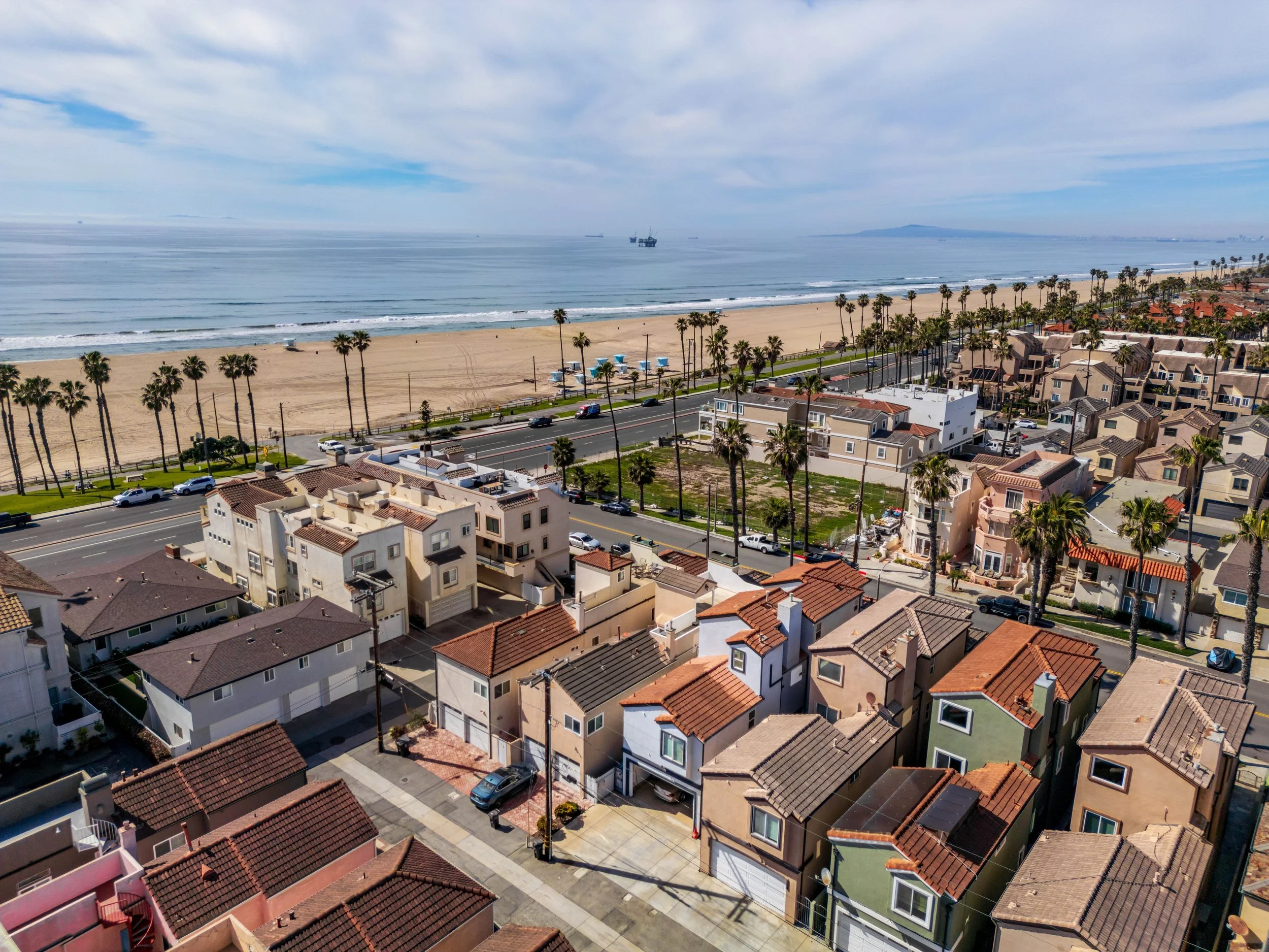 A coastal view showing a sandy beach with palm trees, residential houses with tiled roofs, a road with cars, and the ocean with ships in the distance under a partly cloudy sky.