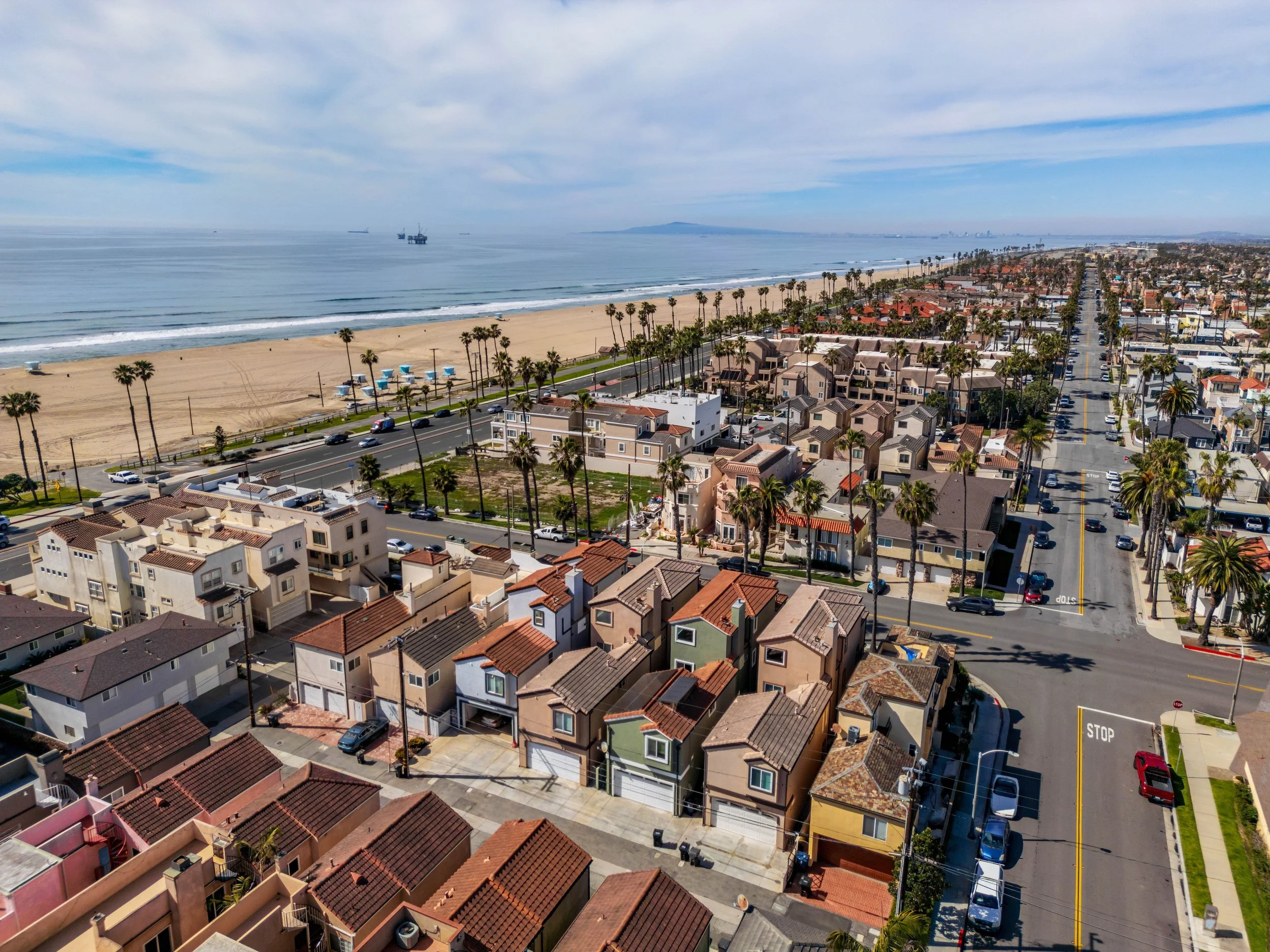 Aerial view of a beachside neighborhood with houses, palm trees, and a wide sandy beach leading to the ocean, with ships sailing in the distance and a cloudy blue sky.