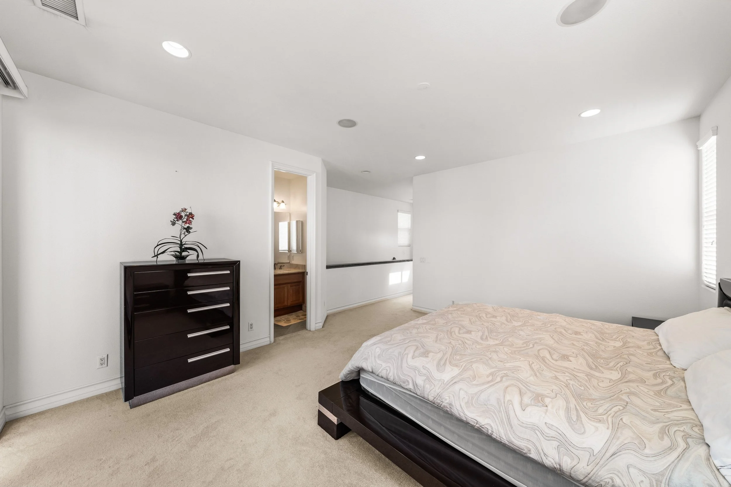 A bedroom with a bed that has a patterned mattress, a black dresser with silver handles and a decorative plant on top, and a doorway leading to a bathroom with a vanity and mirror. The room has white walls, a beige carpet, and ceiling lights.