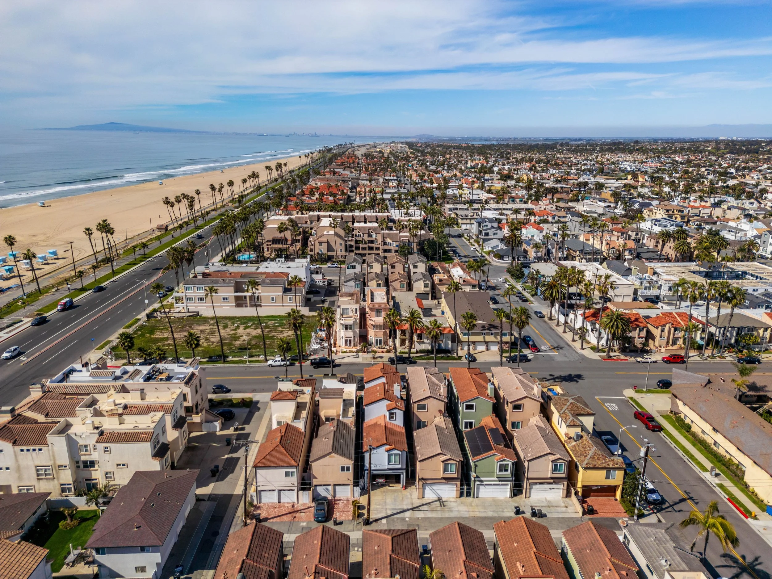 Aerial view of a coastal neighborhood with houses, palm trees, a beach, and the ocean in the background under a partly cloudy sky.