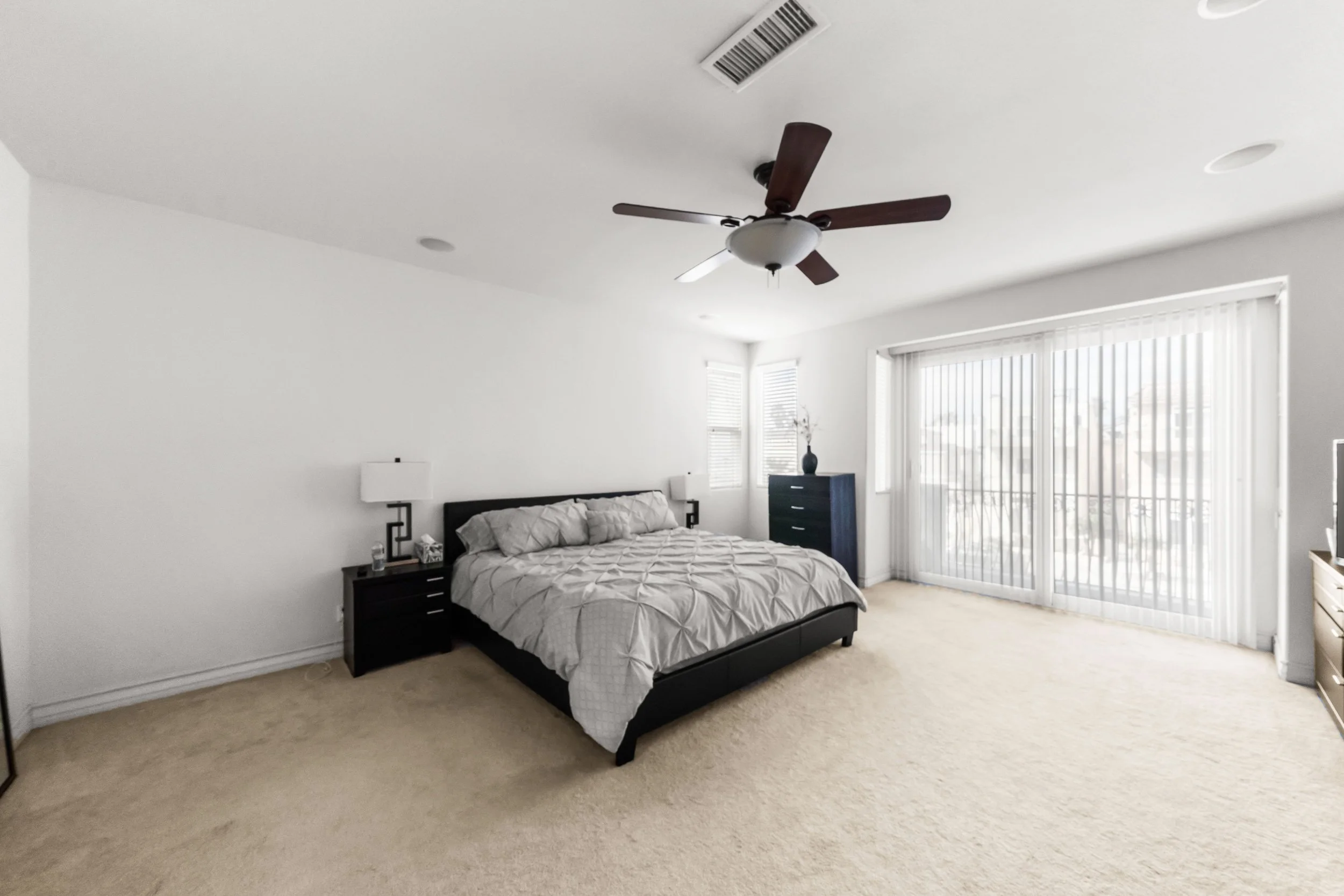 A bedroom with a large bed, black nightstands, and a tall blue dresser near large windows with vertical blinds, a ceiling fan, and white walls.