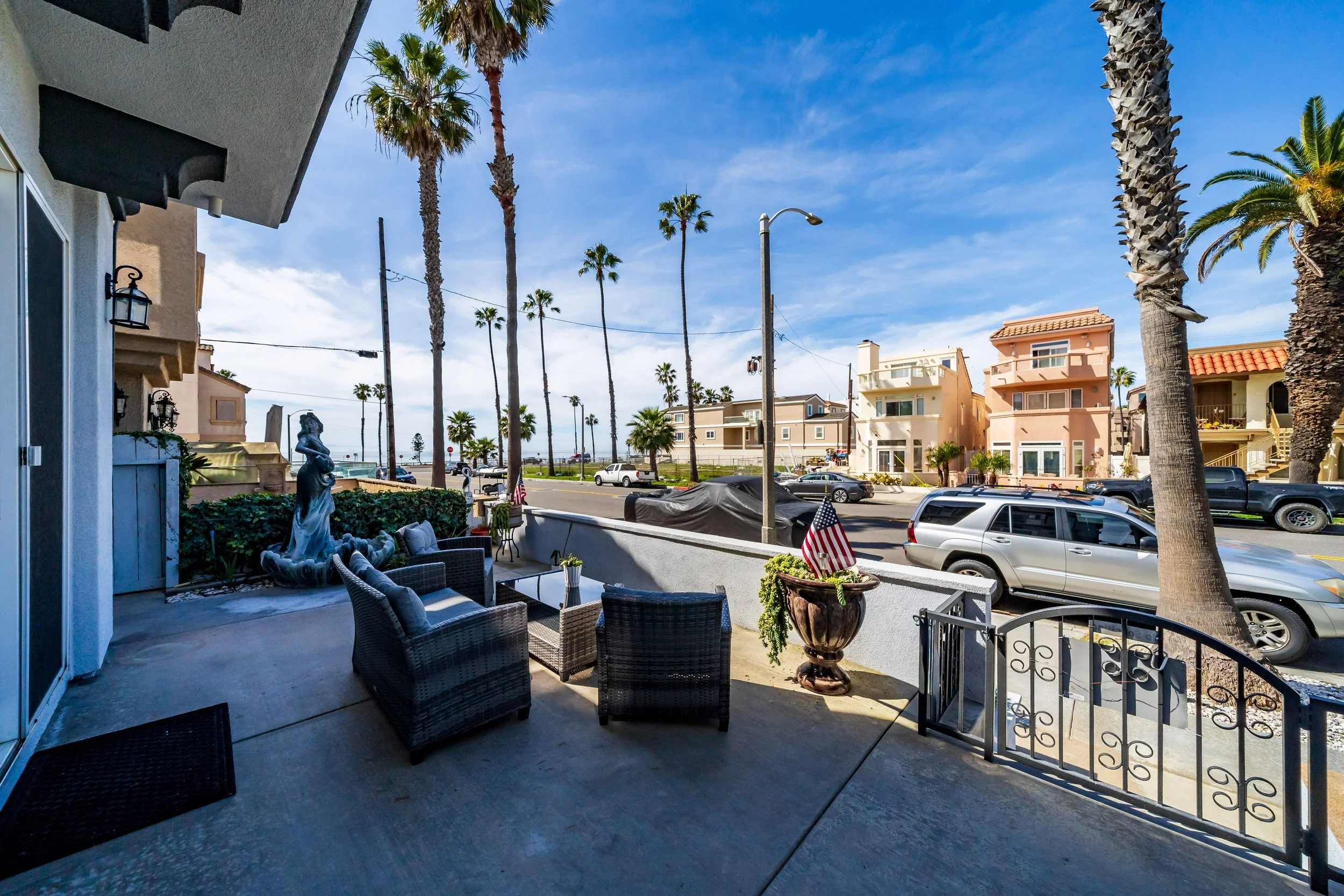 View from a patio showing outdoor seating, a statue, palm trees, and residential buildings under a blue sky.