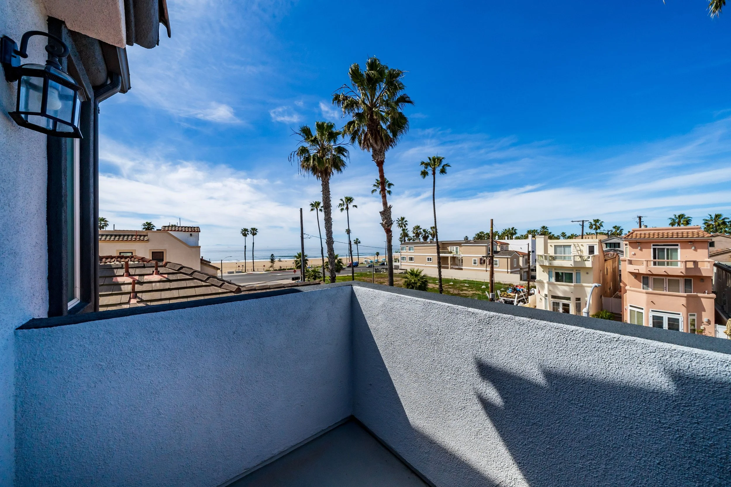 View from a balcony showing palm trees, houses, and a beach in the distance under a blue sky with wispy clouds.