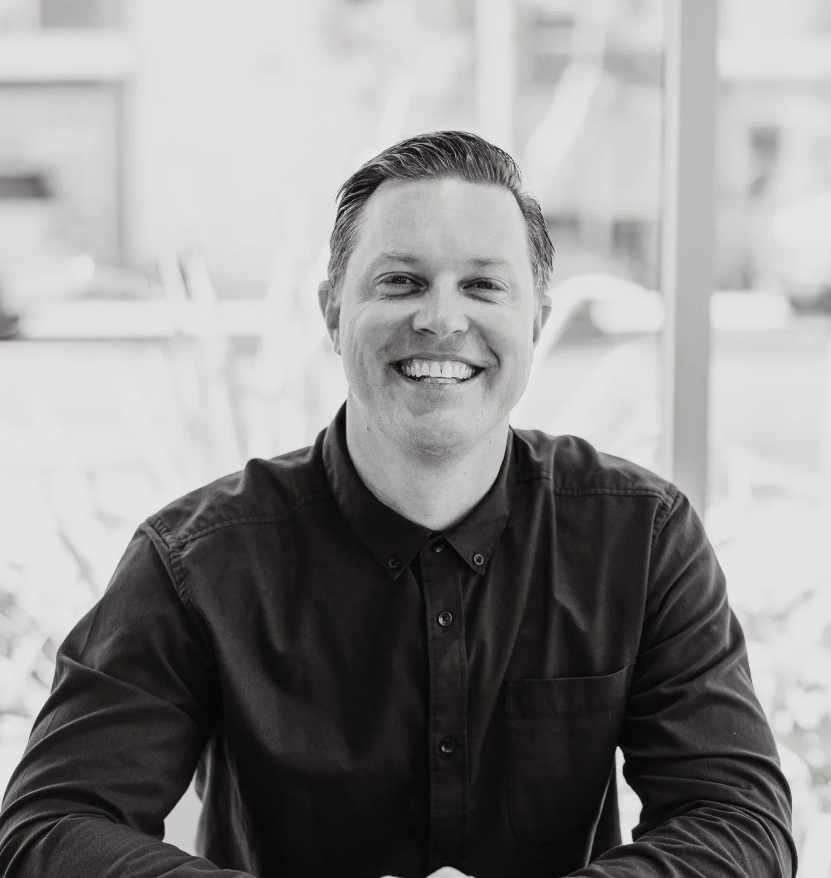 Black and white photo of a man smiling at the camera, seated indoors with shelves in the background.