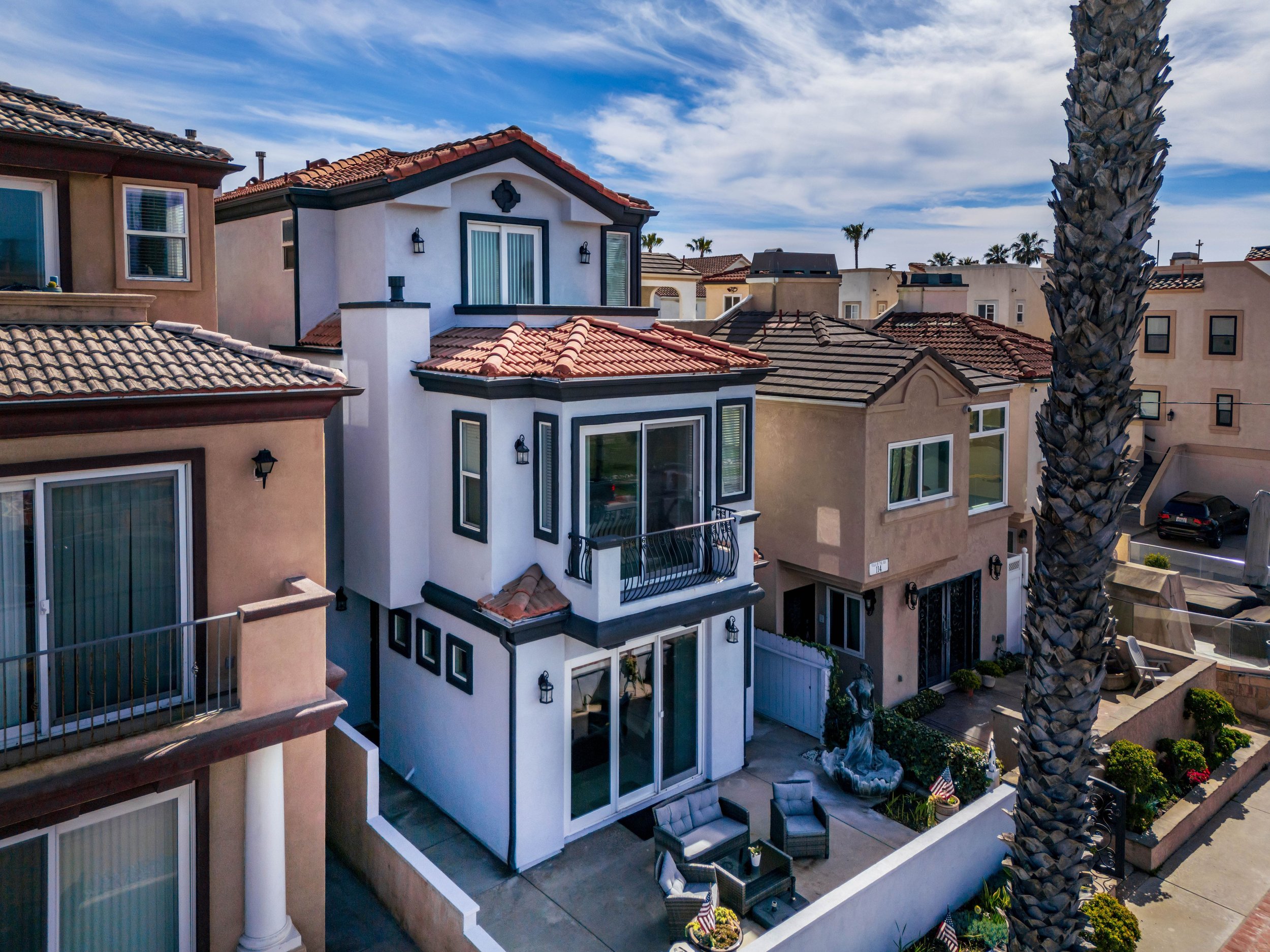 View of multiple houses with tiled roofs, a white house with large windows and a balcony, and palm trees under a partly cloudy sky.