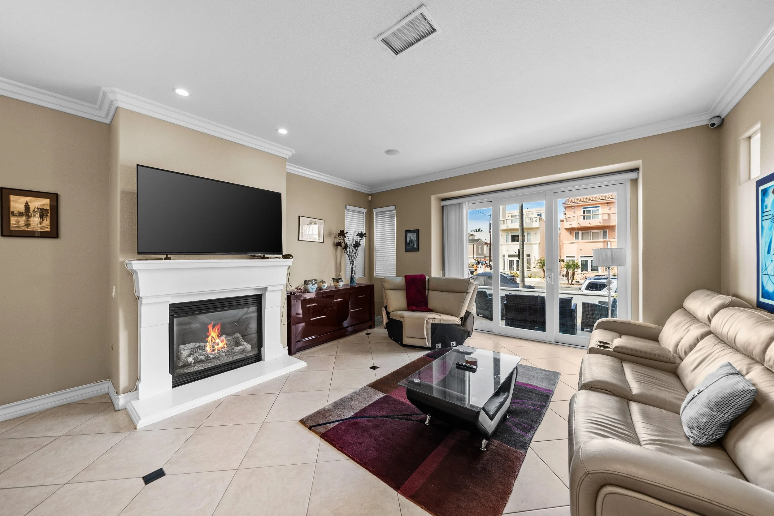 Living room with beige sofas, glass coffee table, fireplace, TV mounted above fireplace, windows with blinds, painting on wall, sliding glass door leading to patio with buildings visible outside.