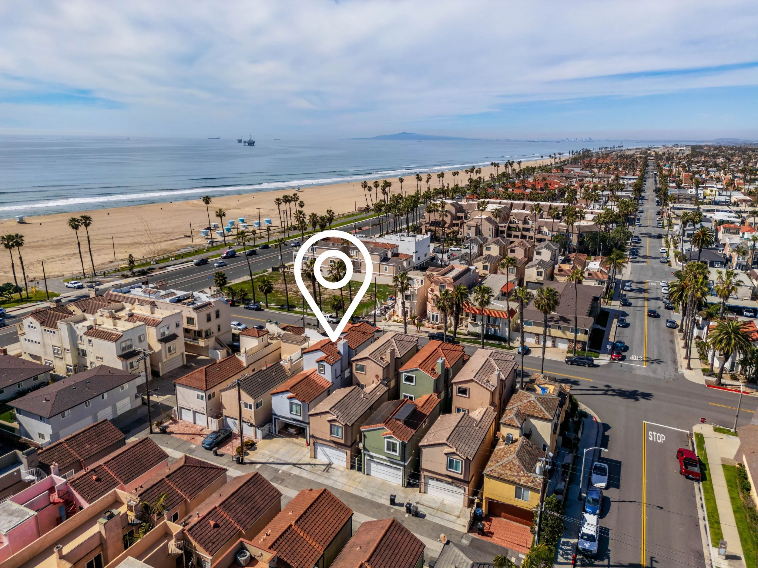 Aerial view of a coastal neighborhood with houses, palm trees, and streets, overlooking the beach and ocean with ships in the distance.