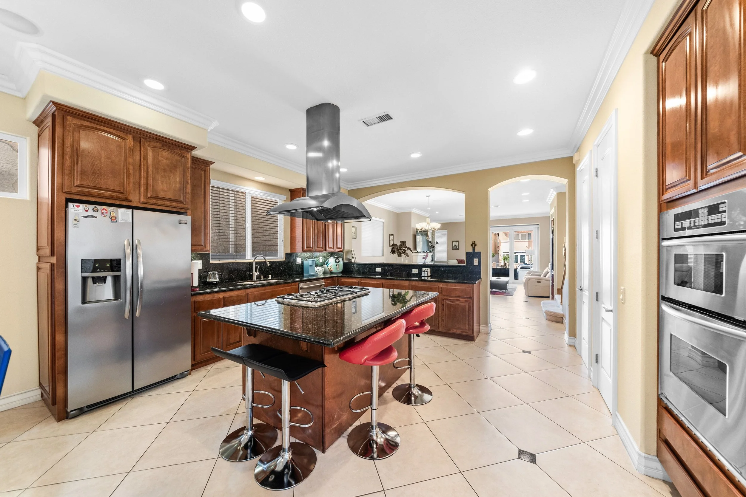 Kitchen with wooden cabinets, stainless steel appliances, black granite countertops, a central island with red bar stools, and a tile floor. A living room is visible through an open doorway.