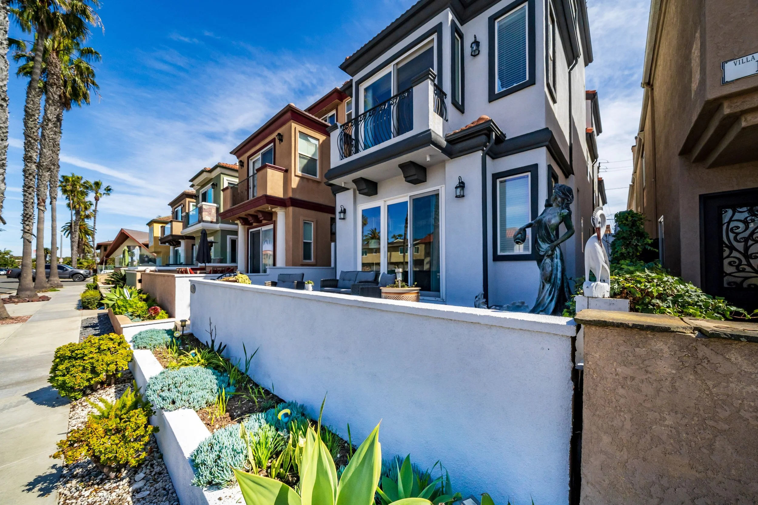 Colorful row of modern houses with balconies, palm trees, and a sidewalk with vibrant plants and flowers in the foreground under a blue sky.