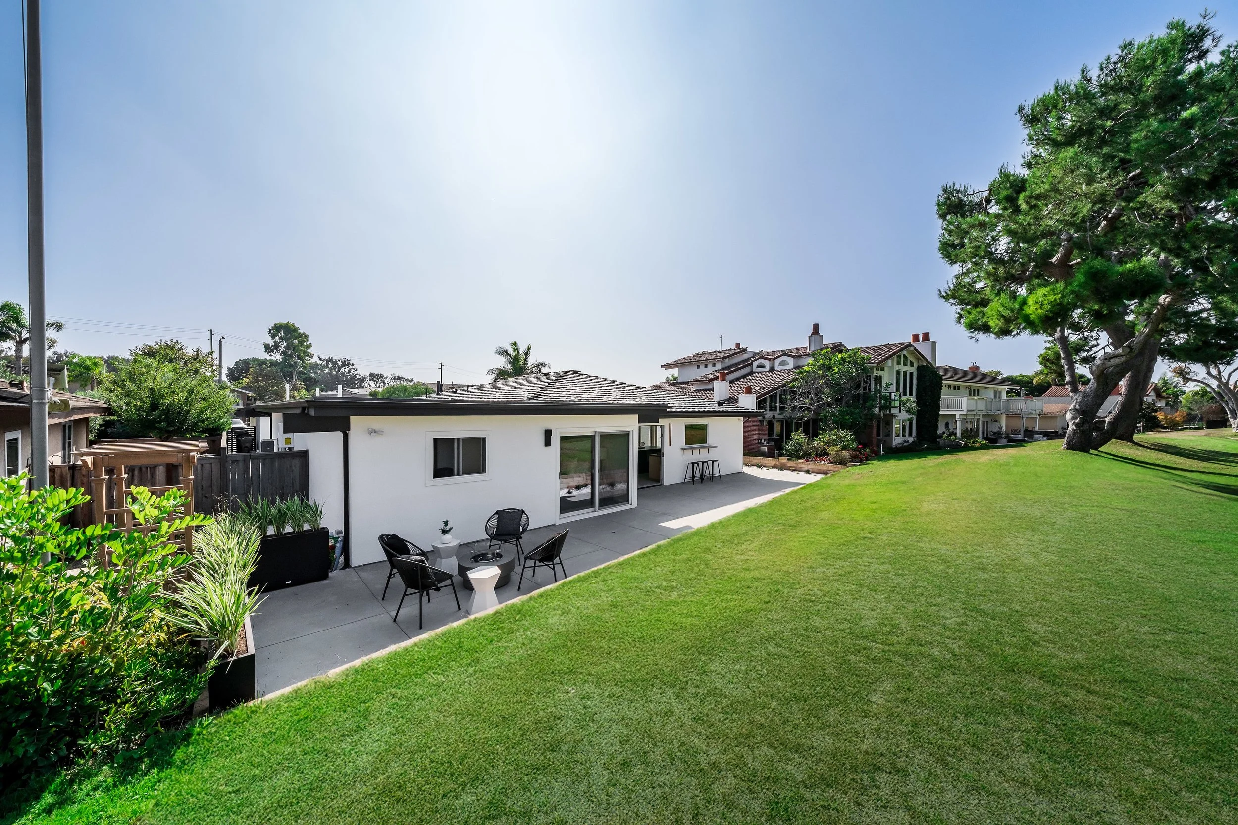 Backyard with green lawn, patio area with outdoor seating, and neighboring houses with trees and clear blue sky.