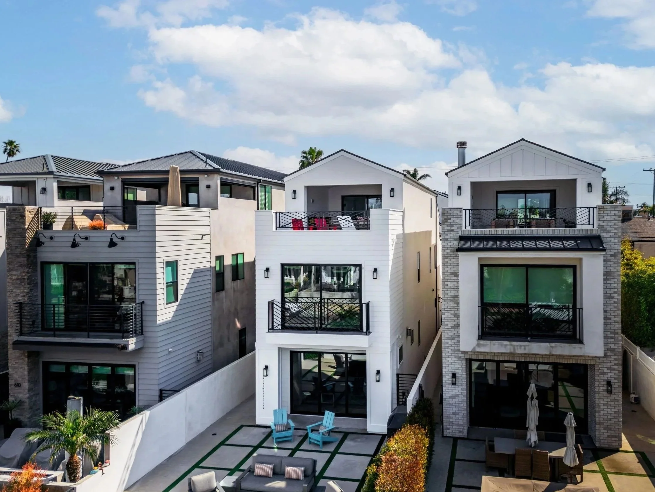A modern three-story house in white with a front living area that has outdoor furniture, trees, and potted plants under a partly cloudy sky.