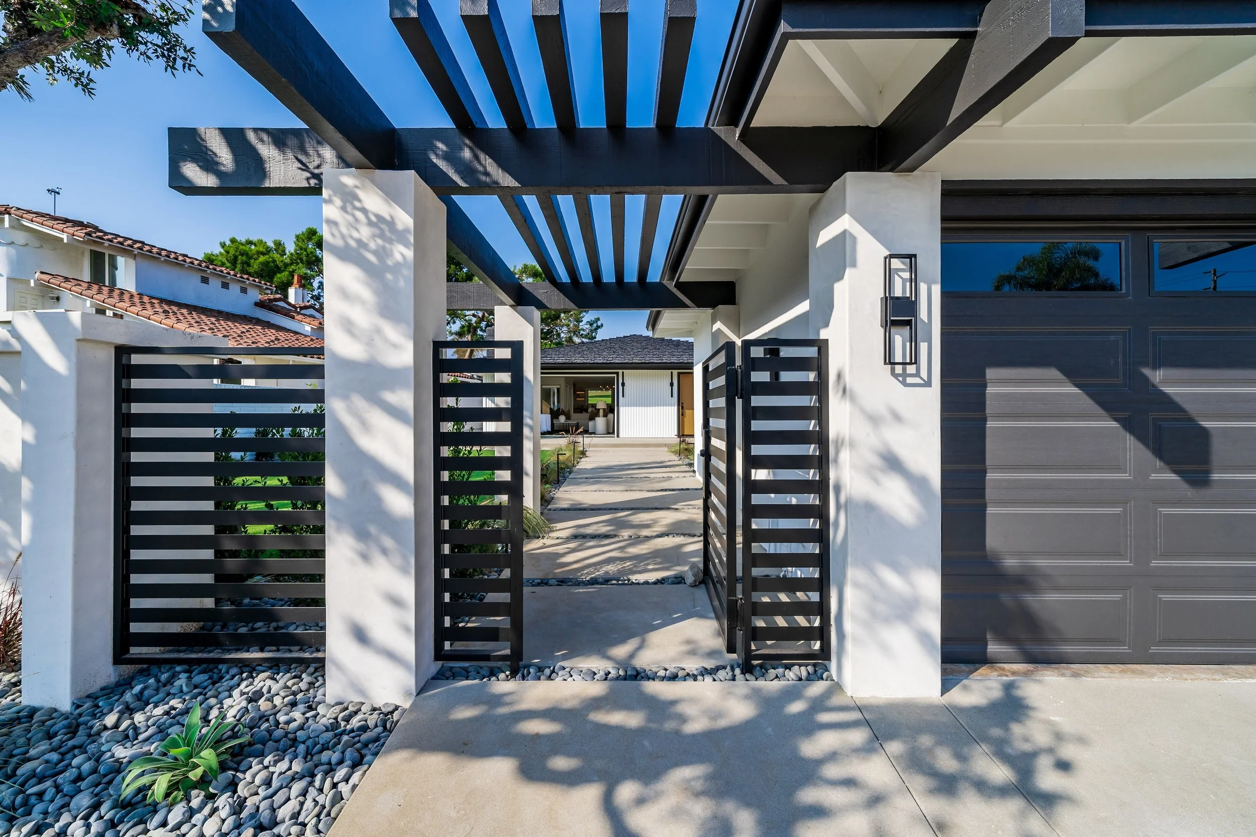A modern house entrance with a black metal gate and horizontal slats, white concrete supports, and a sleek exterior. There is a pathway with rounded stones and plants, with additional buildings visible in the background. The house features large windows and a clean, minimalist design.
