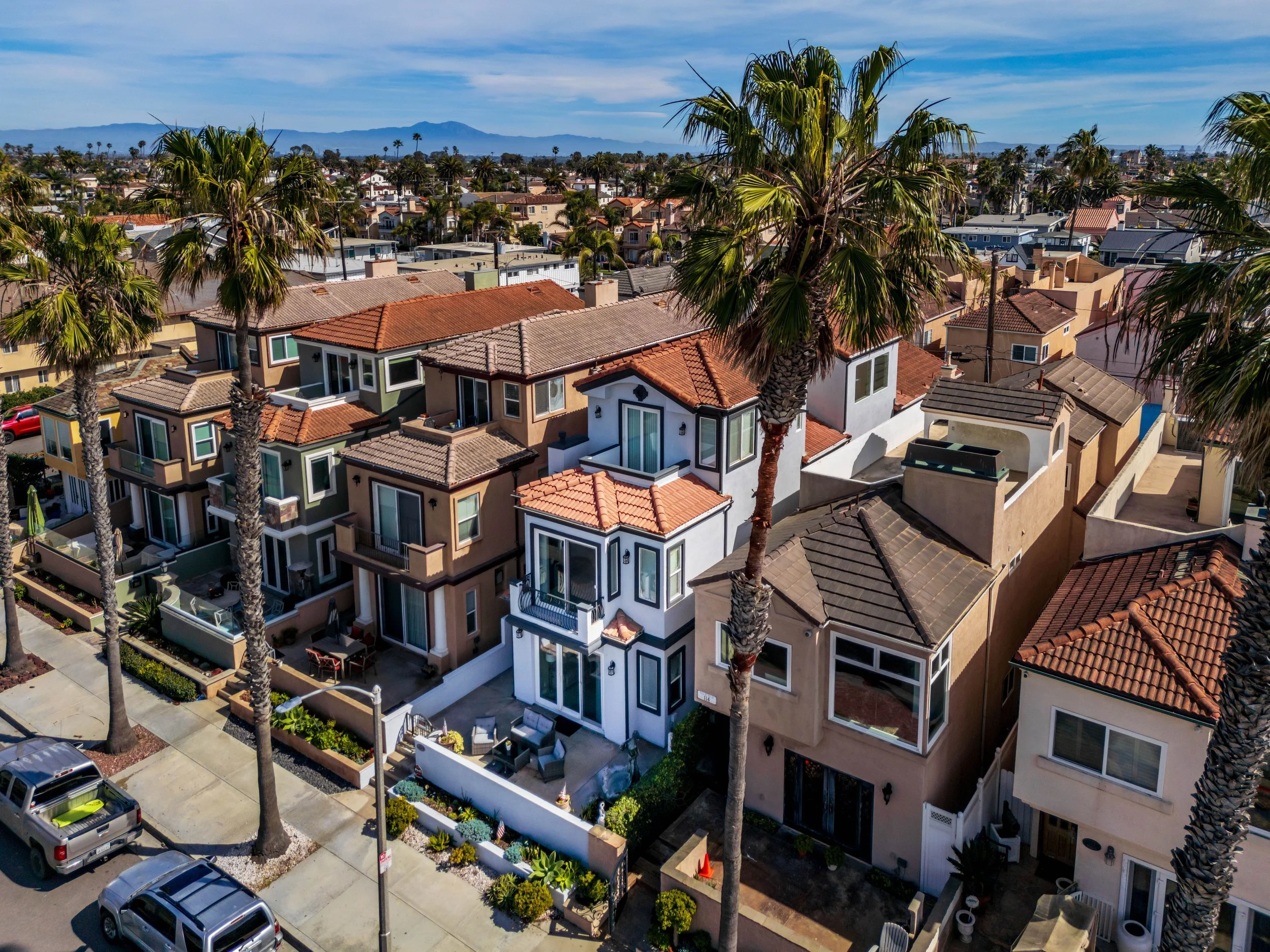 Aerial view of modern residential neighborhood with multi-story houses, palm trees, parked cars, and a distant mountain range.