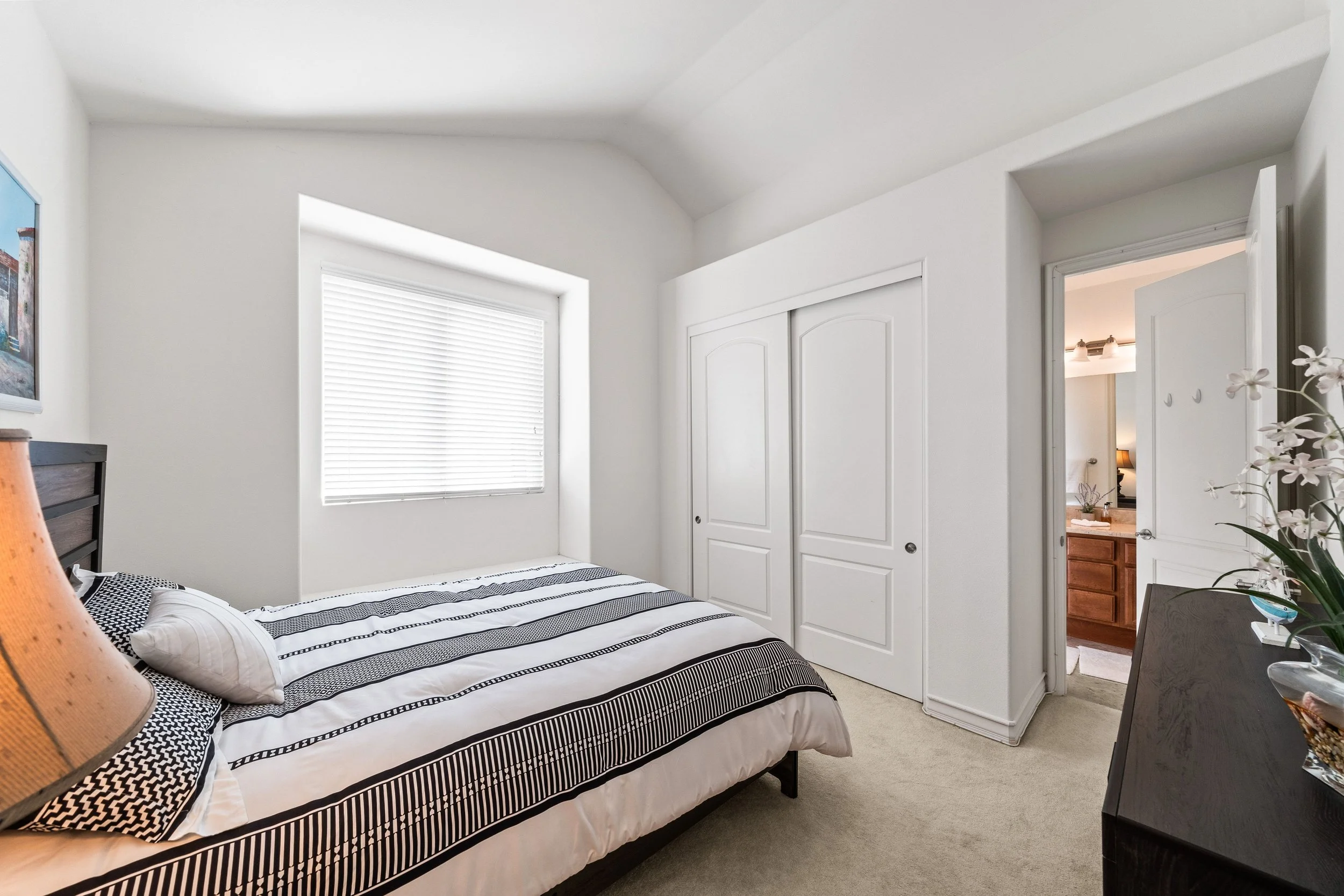 A bedroom with a bed covered in a black and white striped bedspread, a window with white blinds, a closet with white double doors, and a doorway leading to a bathroom with a wooden vanity and a mirror. There is a flower decoration on the nightstand.
