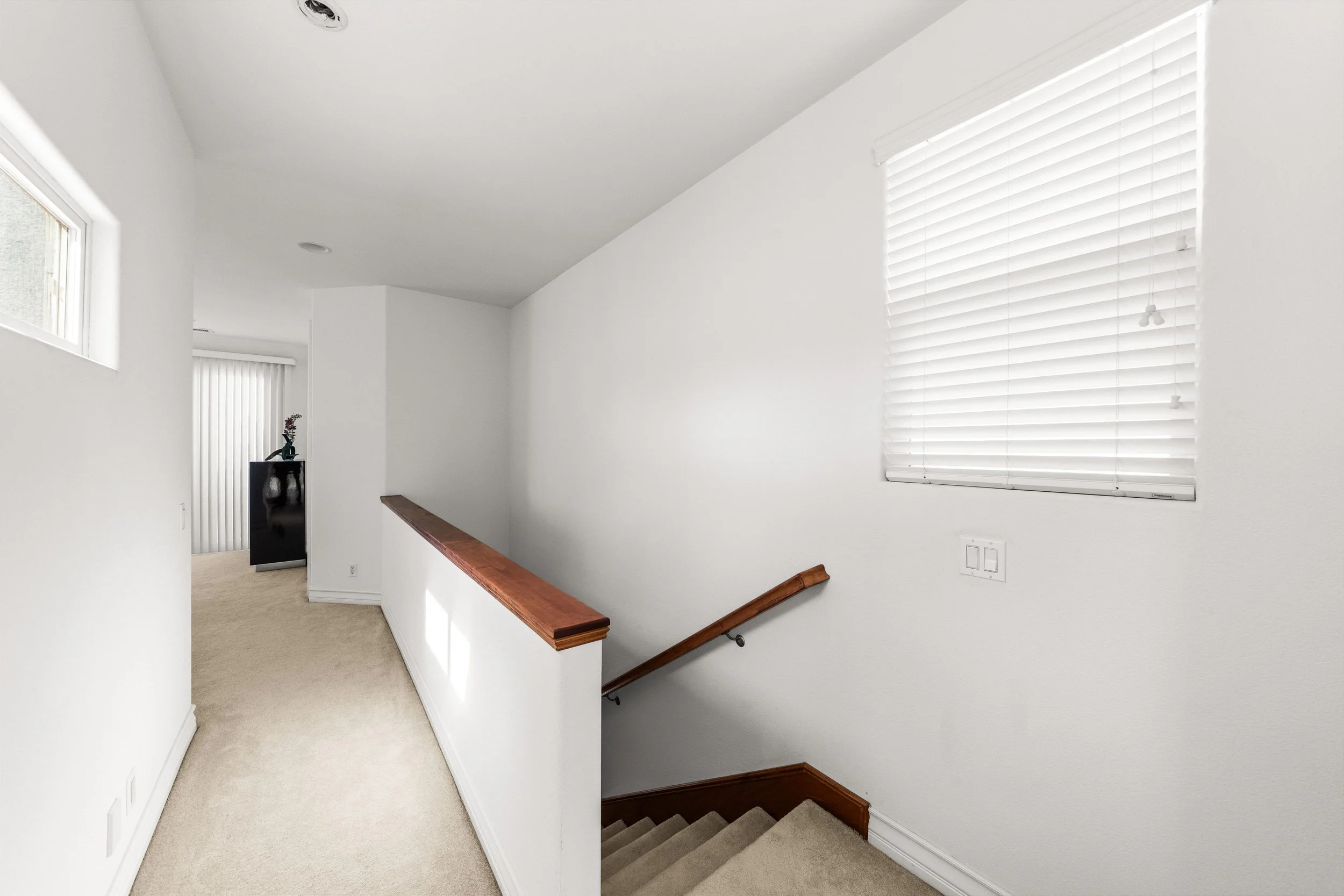 Interior view of a bright staircase landing with white walls, a window with closed blinds, and a wooden handrail along the staircase.